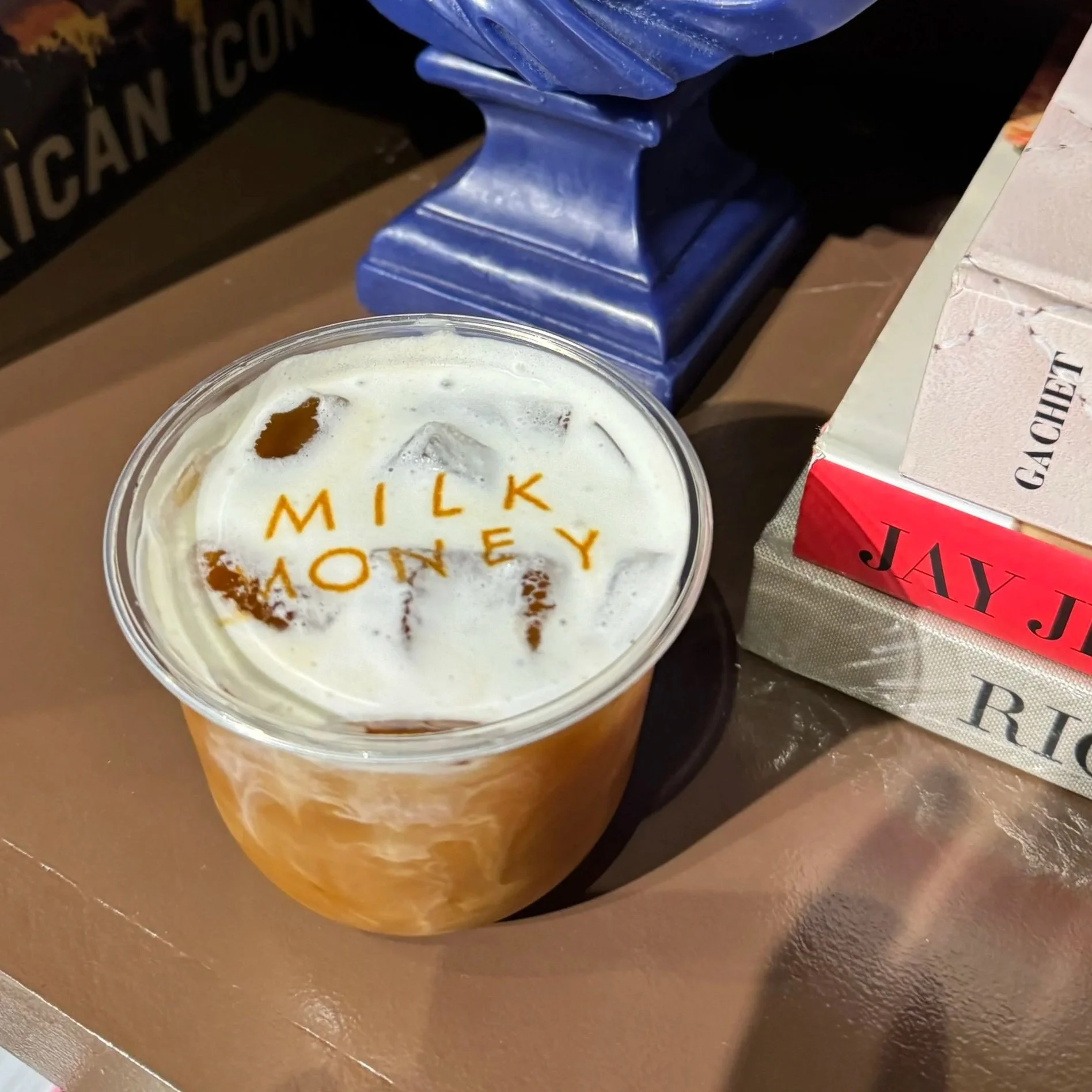 Close-up of a plastic cup filled with caramel iced coffee with a foam top, on a brown table, with part of a box and a blue and black object in the background.