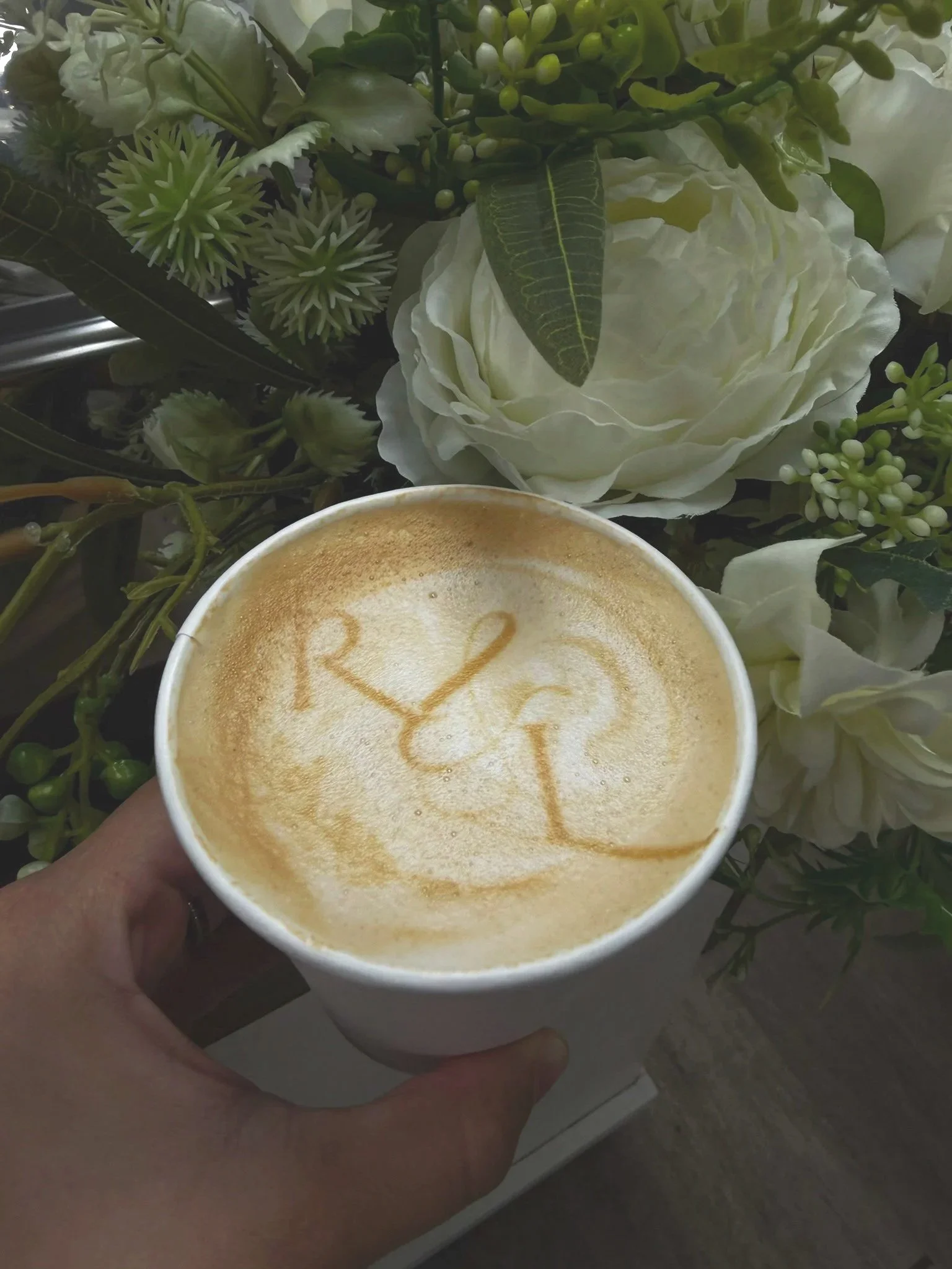 A cup of coffee with foam art that reads 'Happy 21' and a background of white and green artificial flowers.