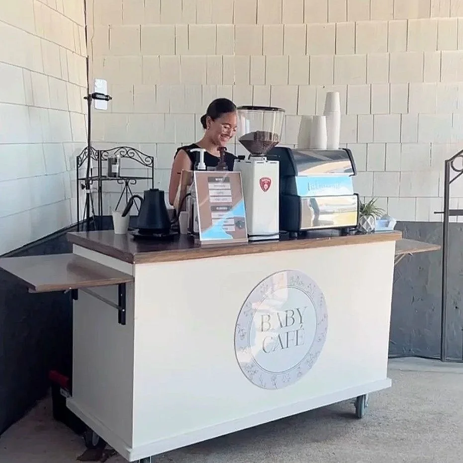 A woman behind a small coffee stand with a sign that reads 'Baby Cafe', smiling, with coffee equipment on the counter and a beige brick wall in the background.