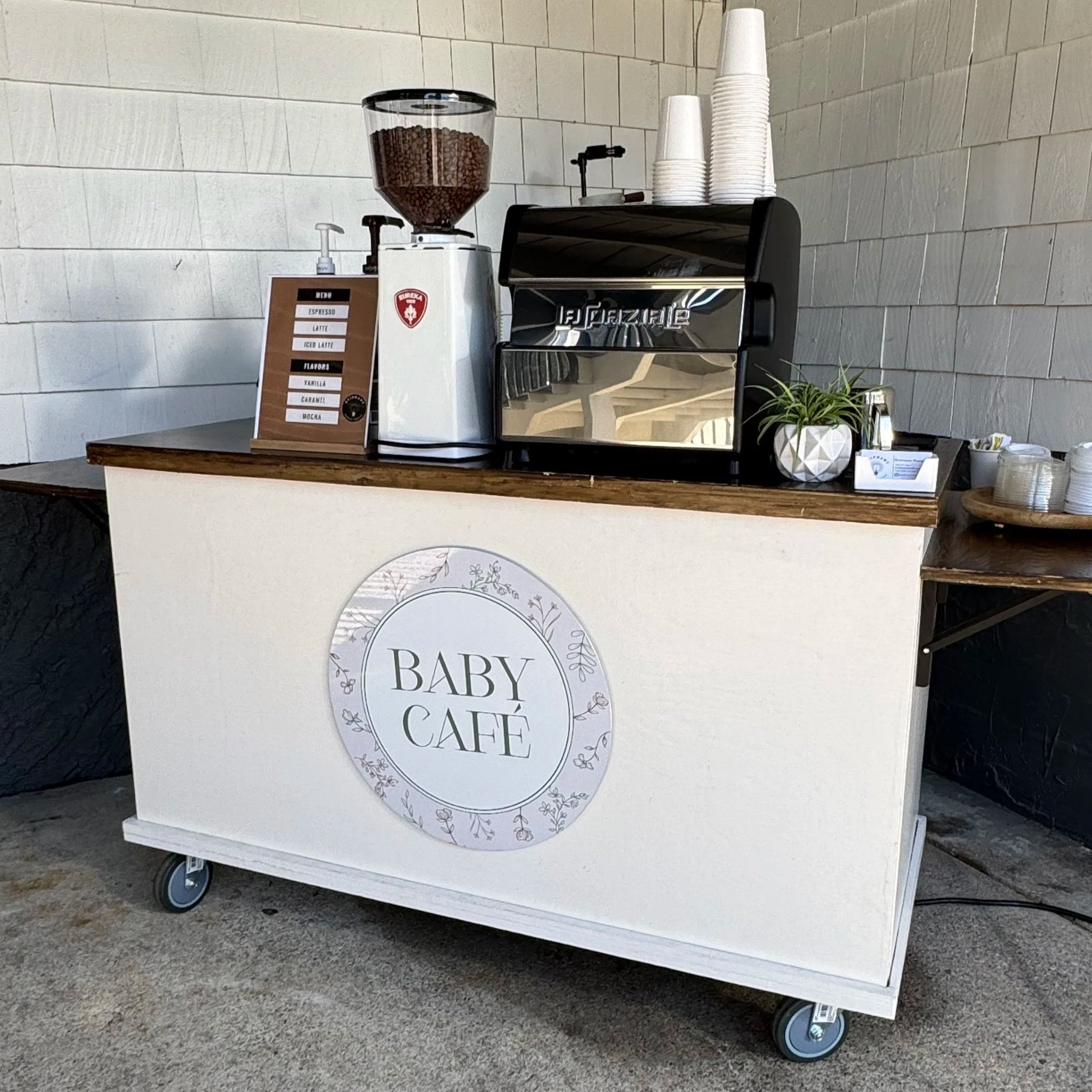 A coffee stand on wheels with a La Marzocco espresso machine, disposable cups stacked on top, a coffee bean grinder, and a menu sign with flavors such as vanilla, caramel, and mocha in an outdoor setting.
