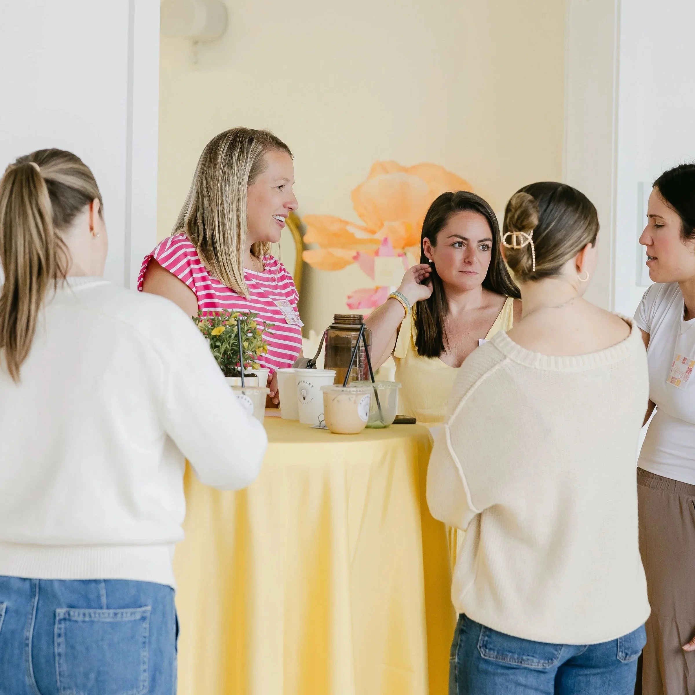 Group of women talking at a table with drinks, against a yellow wall with floral artwork in the background.