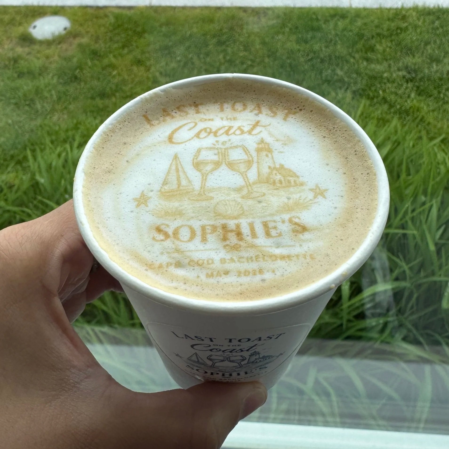 A person holding a white coffee cup with foam art that has a beach scene, including a lighthouse, starfish, and waves, with the text 'Last Toast' written on the cup.