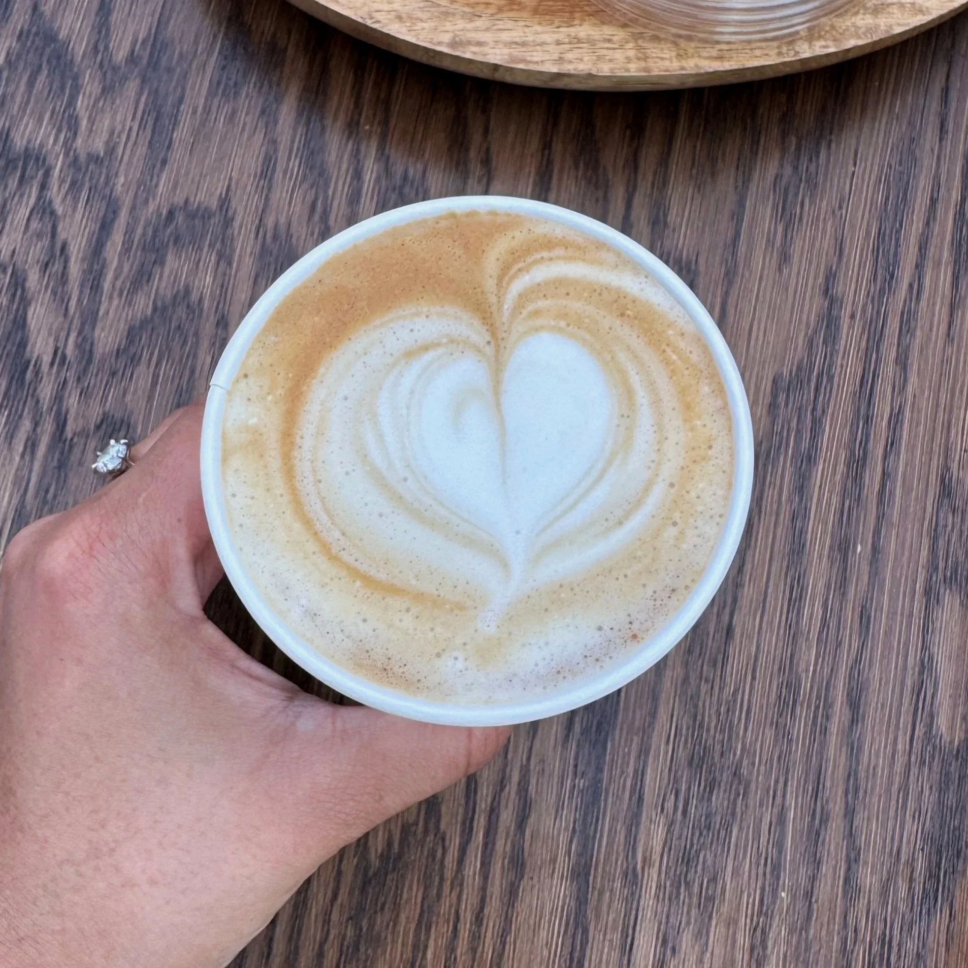A person holding a paper cup of coffee with latte art on top, featuring a heart shape design. The background shows a wooden surface and a round wooden tray.