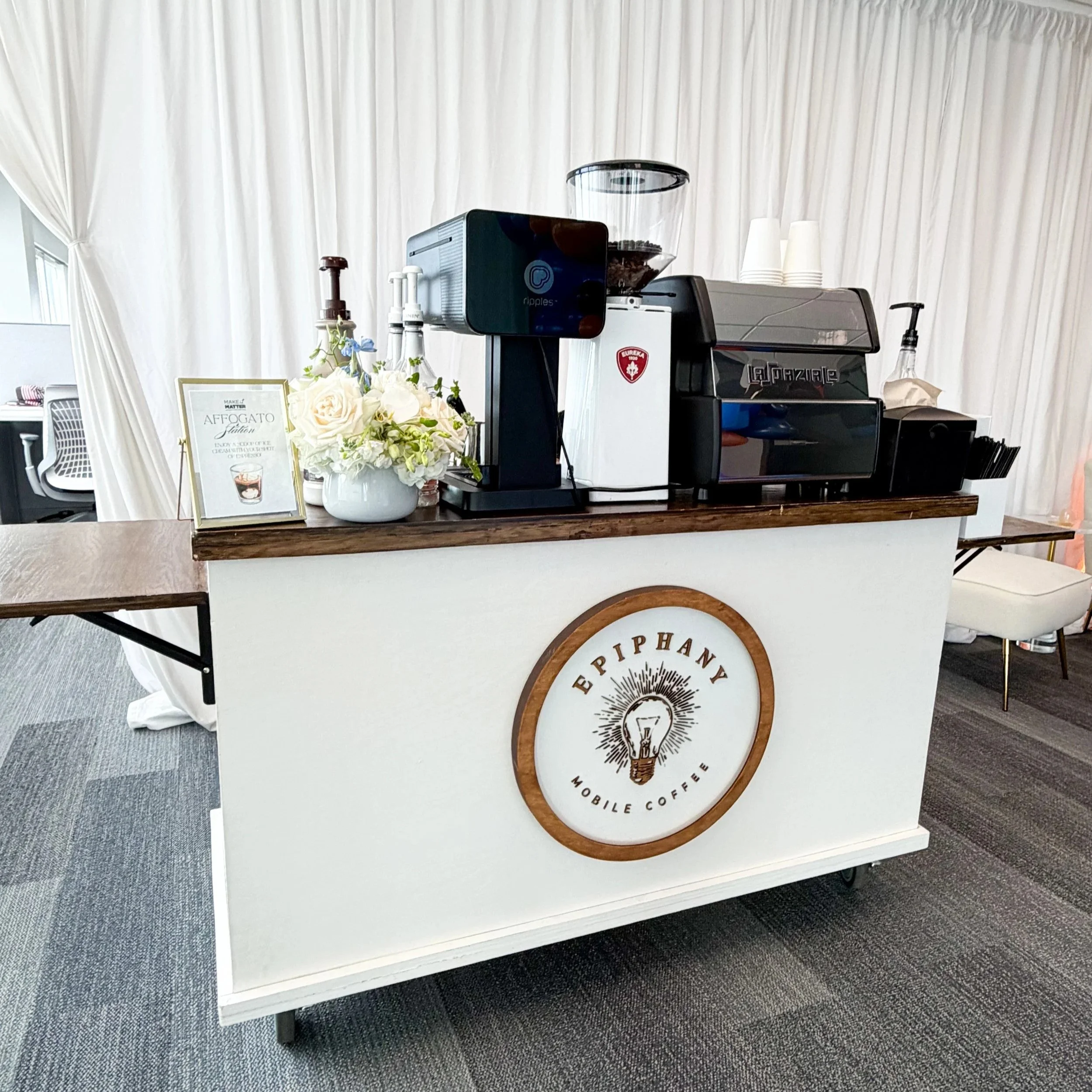 A white coffee bar with a large round logo that reads 'Epiphanie Mobile Coffee,' a coffee machine, a container of coffee grounds, a small bouquet of white and purple flowers, and a framed sign promoting affogato.