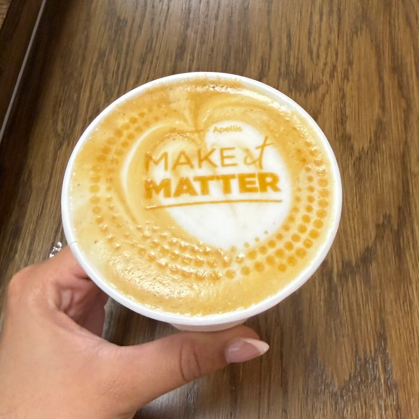 A hand holding a paper cup with a latte art design and the words 'Make It Matter' on the foam, placed on a wooden surface.