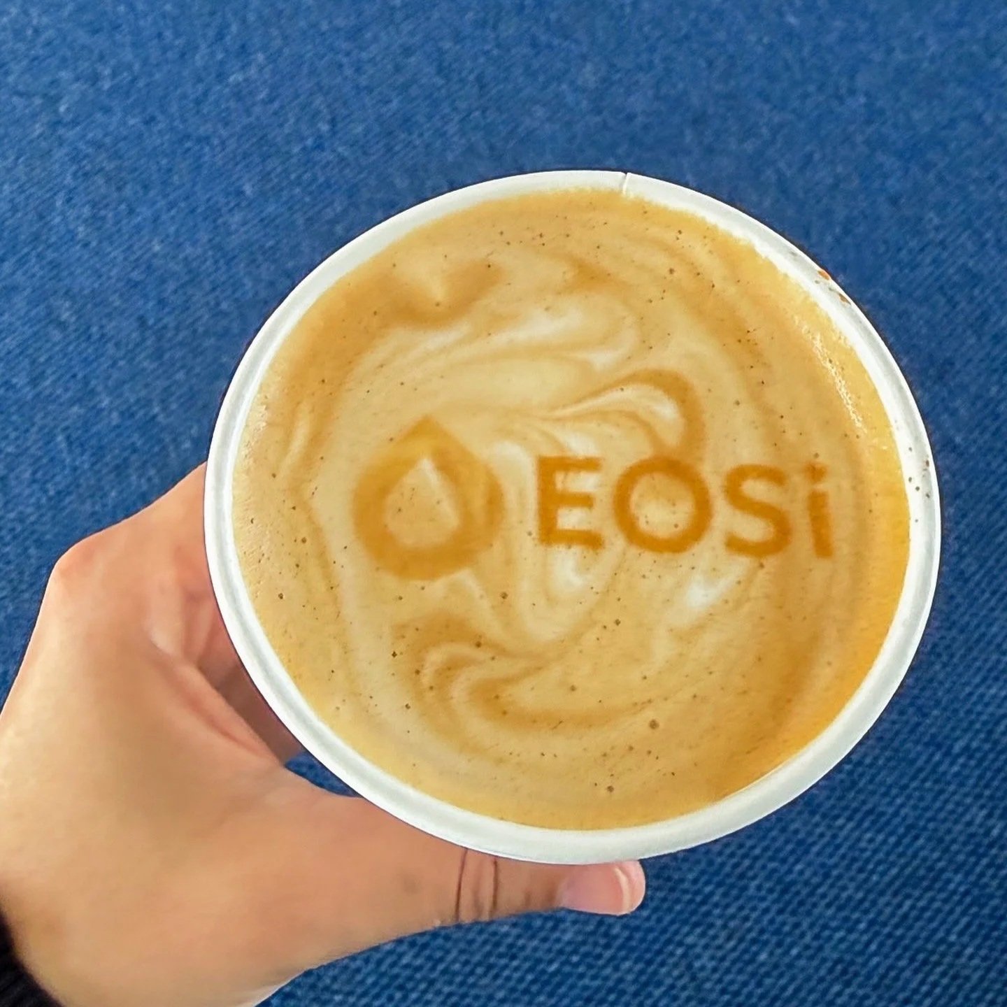 Hand holding a coffee cup with the 'EOSI' logo visible on the surface of the coffee.