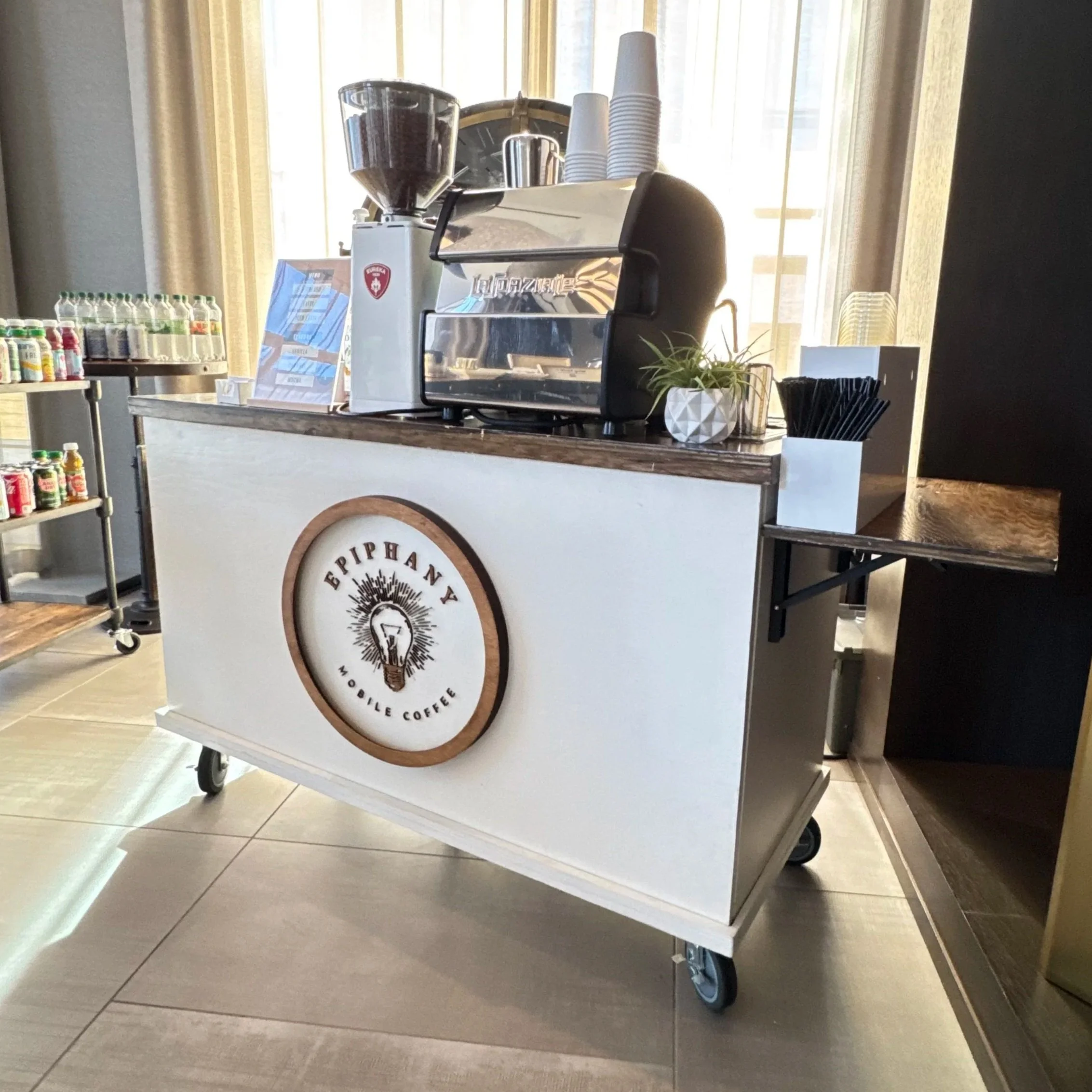 A mobile coffee cart with a coffee machine, disposable cups, and bottled drinks on a table, with a window and curtains in the background.