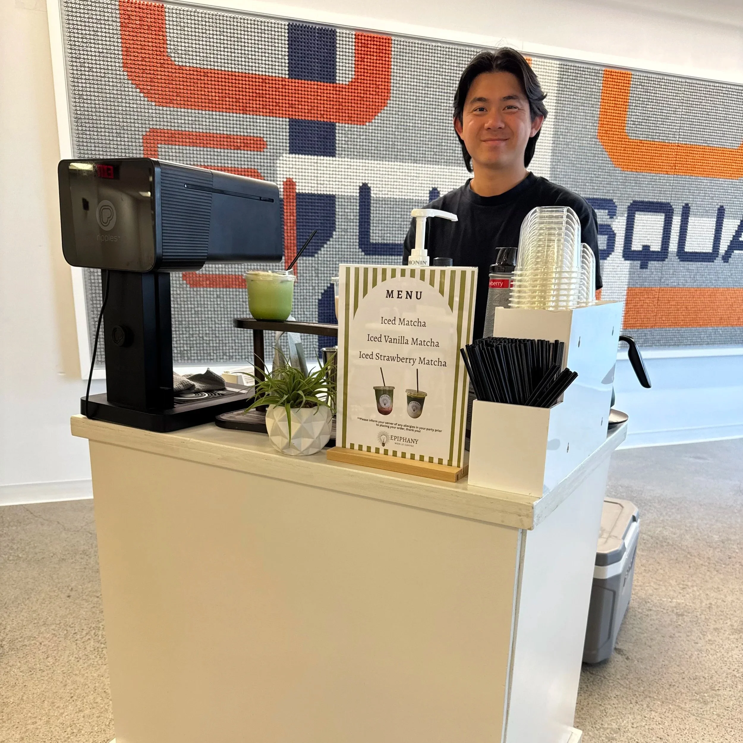 A man with black hair smiling behind a coffee shop counter with a menu sign and a plant. In the background, a large colorful LED display with abstract designs.