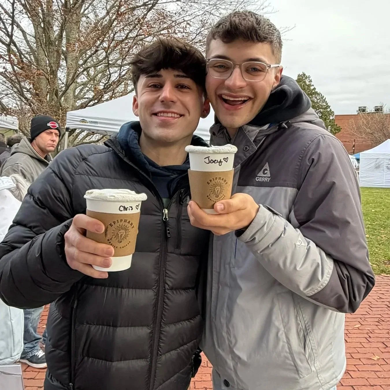 Two young men smiling and holding cups of coffee with handwritten names on the cups. They are outdoors on a cloudy day, dressed in jackets, with trees and tents in the background.