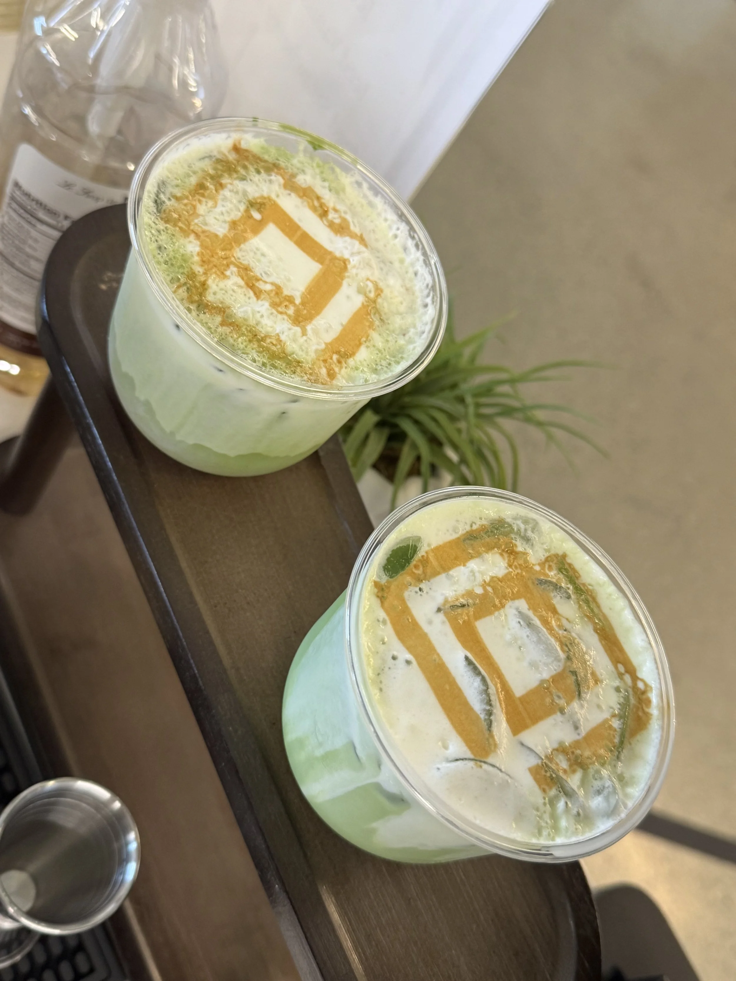 Two green beverages with ice in clear glasses, topped with whipped cream and orange syrup, sitting on a wooden surface. A small plant is visible in the background.