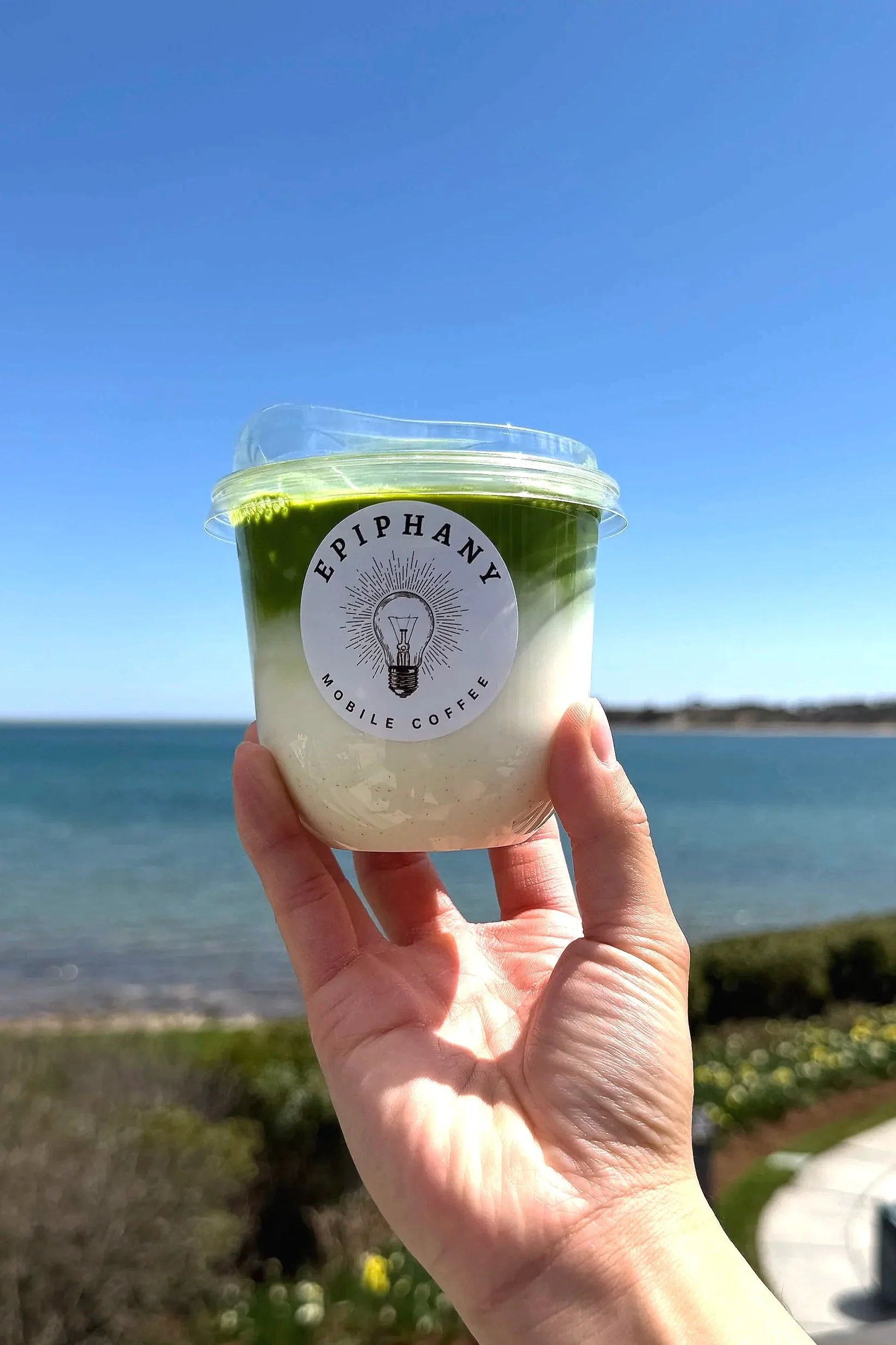 A hand holding a cup of green and white layered beverage outdoors with an ocean and blue sky in the background.