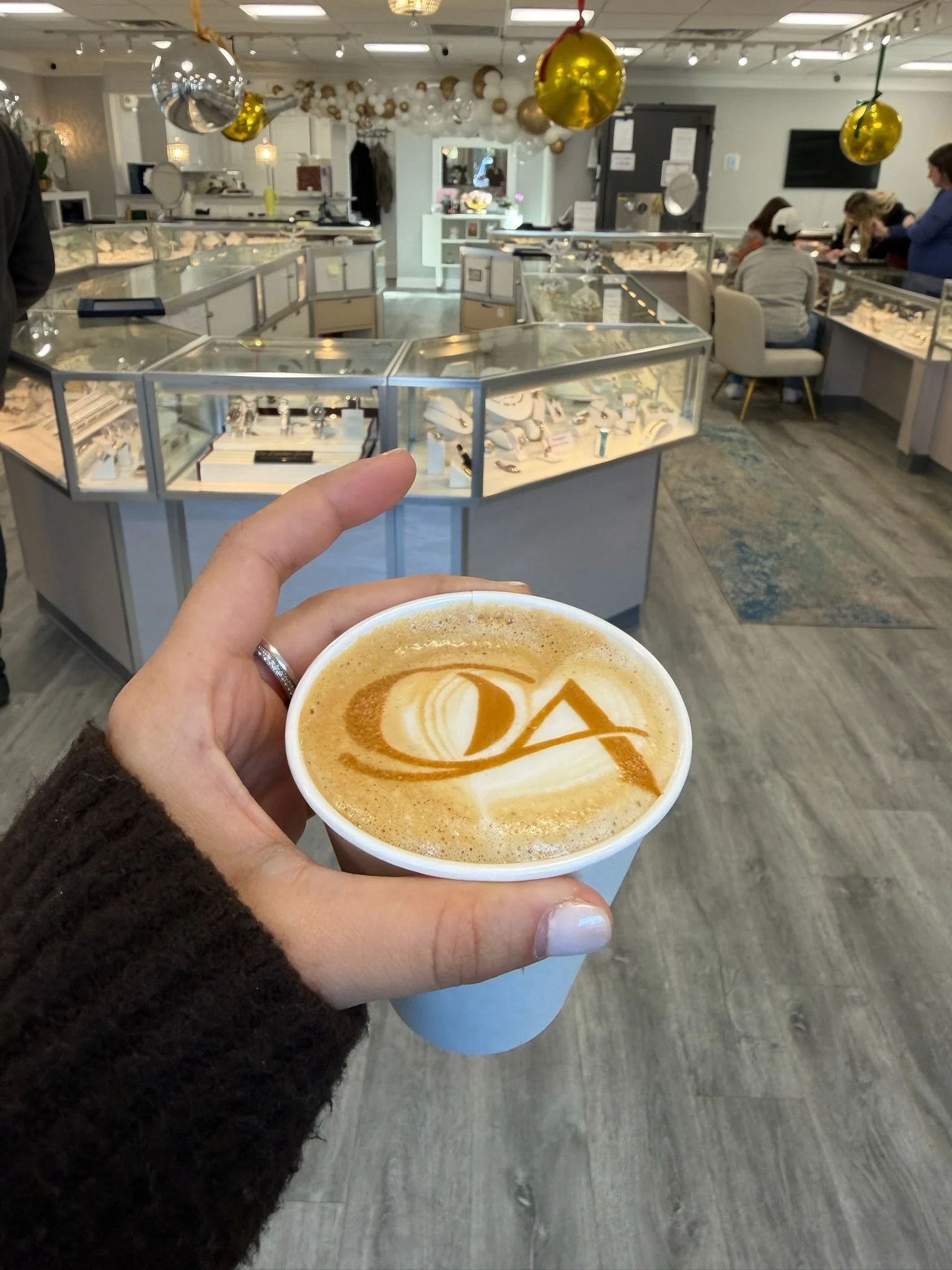 Hand holding a coffee cup with latte art spelling 'QA' inside a jewelry store decorated with gold and silver balloons.