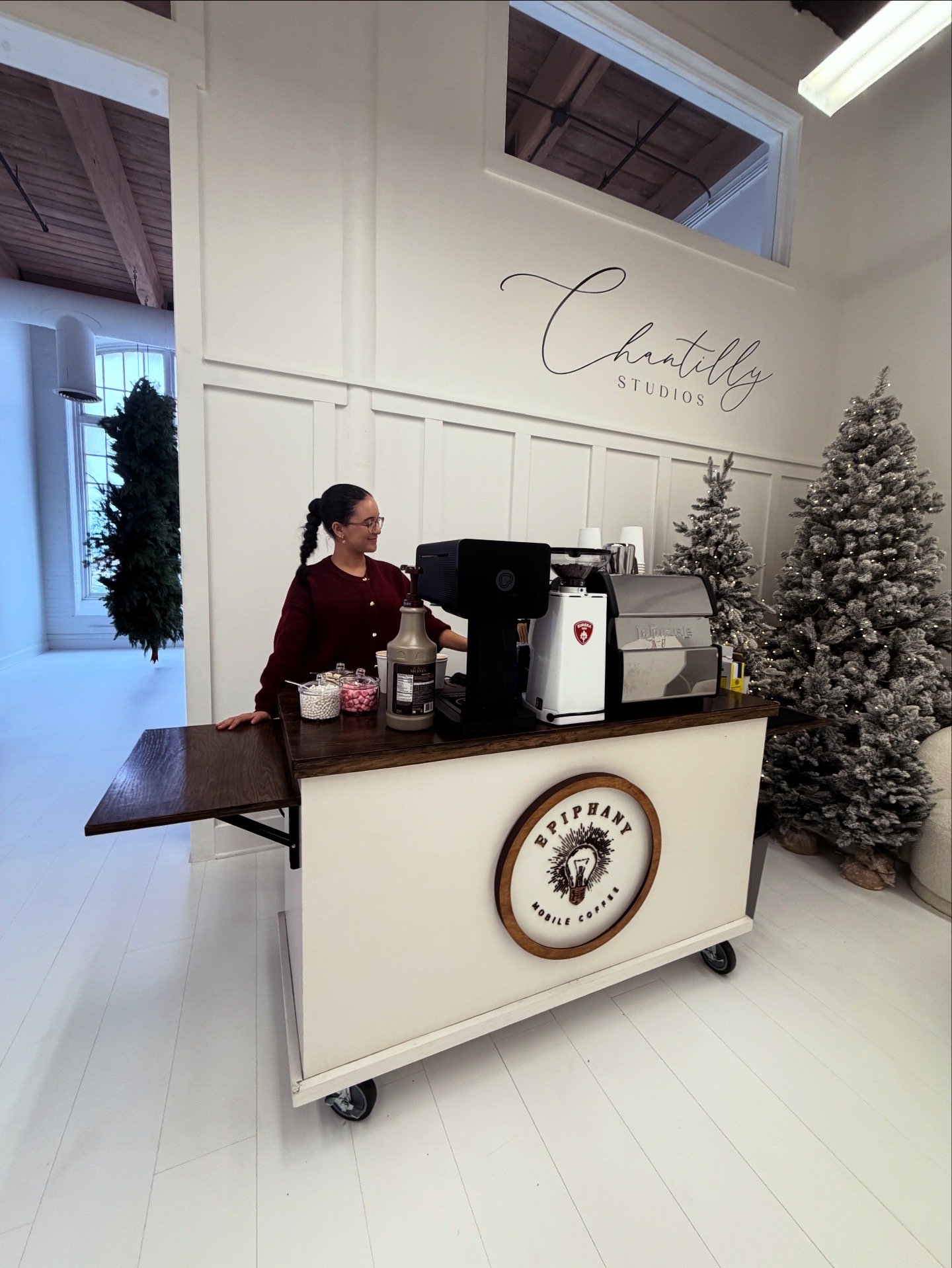 A woman working at a mobile coffee cart inside a studio named 'Chantilly Studios'. The cart has a logo that says 'Ebpiry' with a lightbulb illustration. There are Christmas trees and decorations in the background.