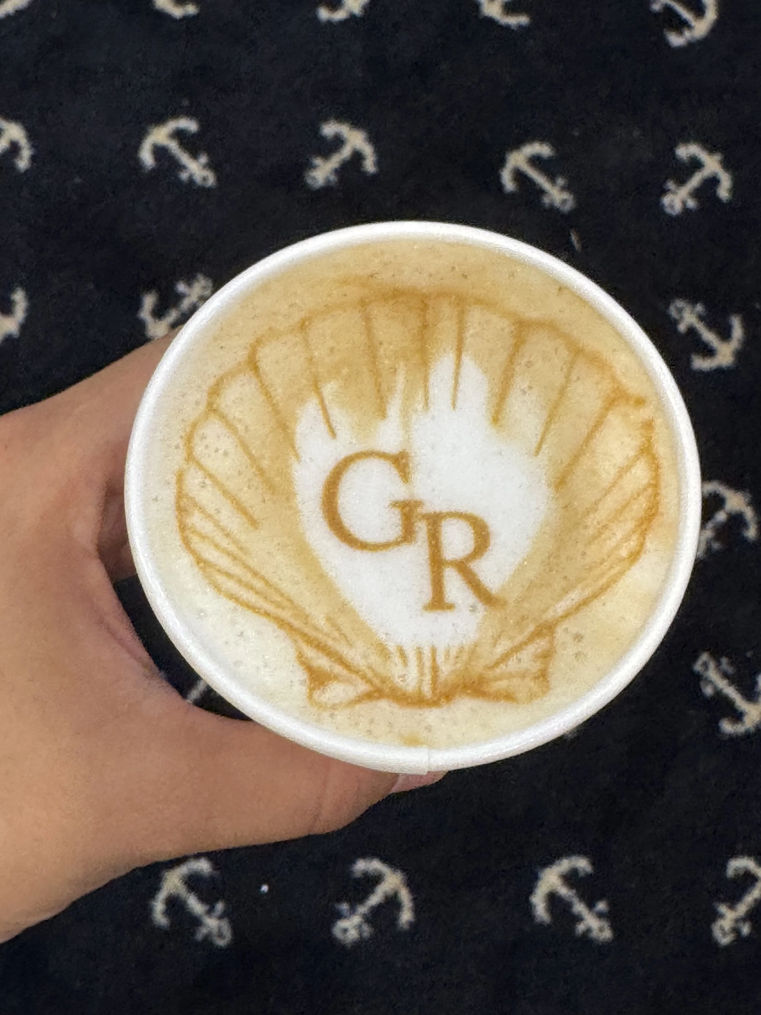 A cup of coffee with latte art in the shape of a lemon slice and the letters 'G R' in the center, held in a person's hand against a fabric background with white anchors.
