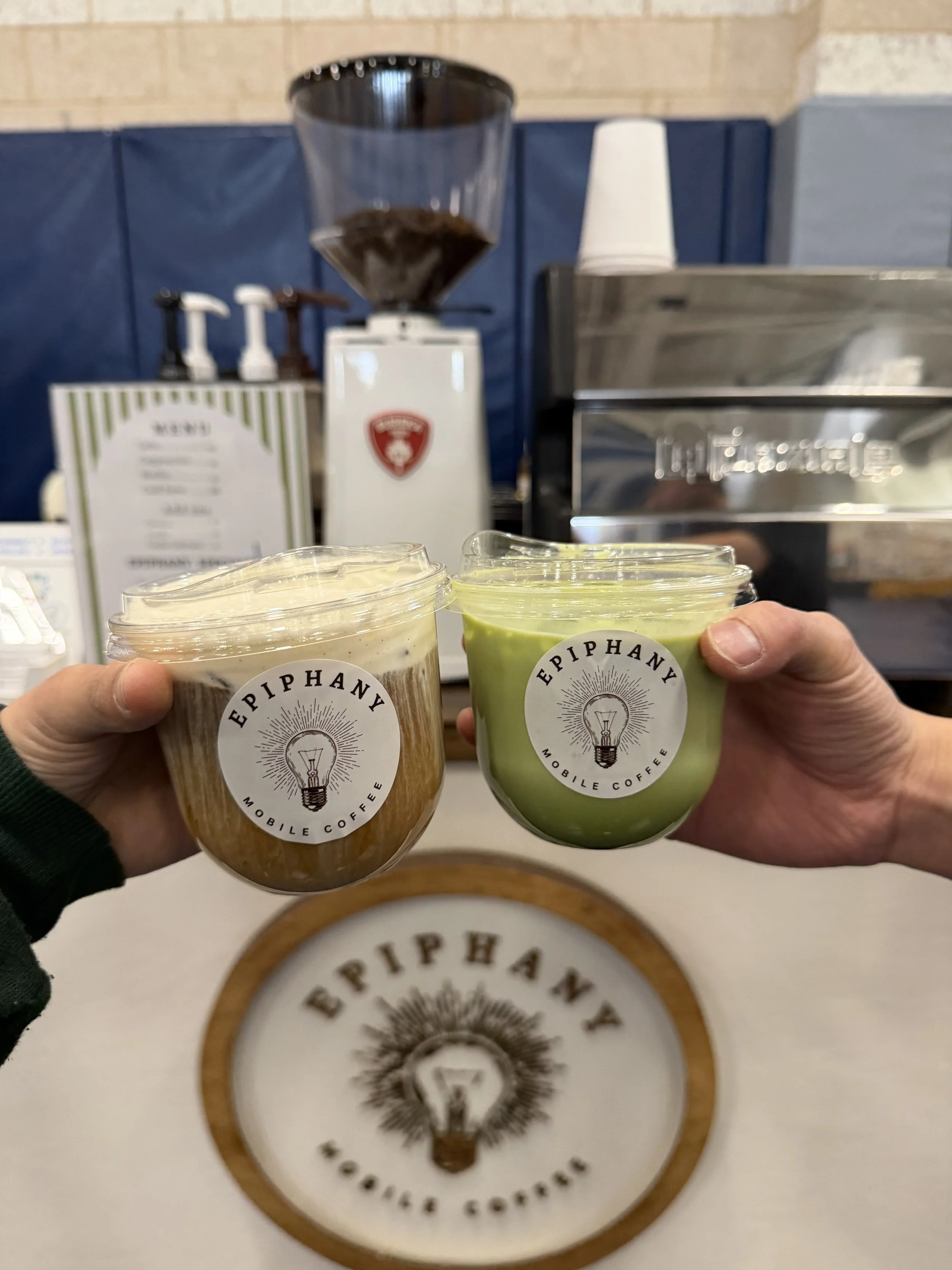 Two cups of coffee from Epiphany Mobile Coffee being held in front of a coffee shop counter with a coffee grinder and coffee syrups in the background.