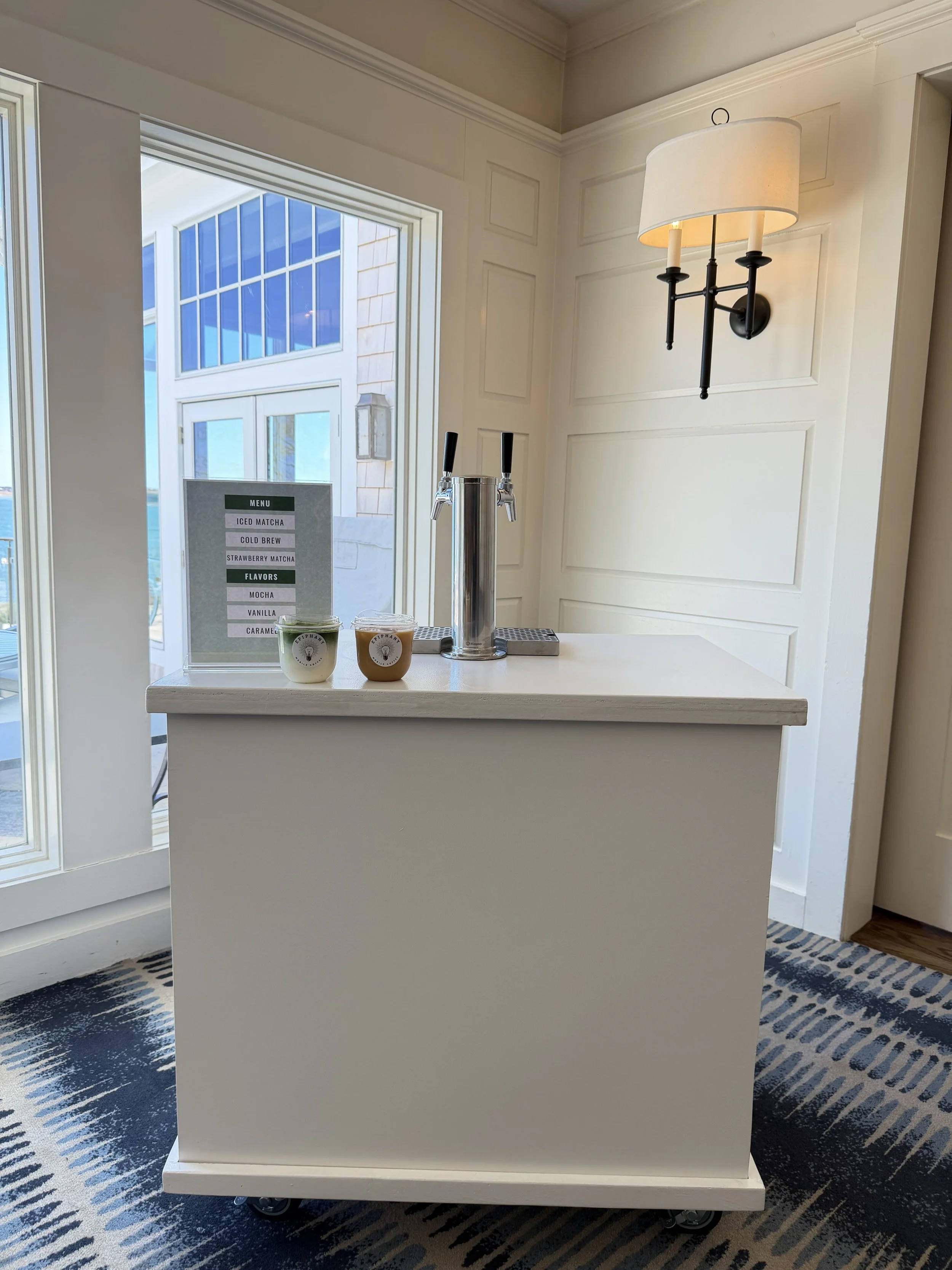 A white counter with a beer tap on top, positioned in front of a window with a view of a building exterior, and a menu sign featuring ice matcha, cold brew, strawberry matcha, and flavors like mocha, vanilla, and caramel; also a wall-mounted light fixture with a white lampshade behind the counter.