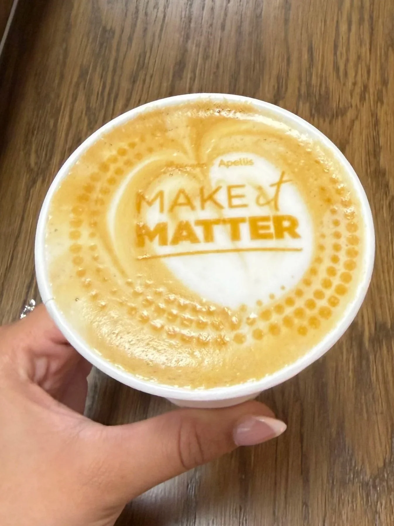 A hand holding a cup of latte with a latte art design that says 'Make It Matter' and the word 'Apellus' above it, on a wooden surface.