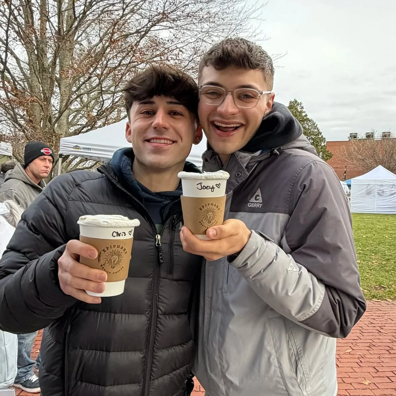 Two smiling young men holding cups of coffee or hot chocolate outdoors on a cloudy day, with a man in a beanie and jacket standing in the background near a white tent.