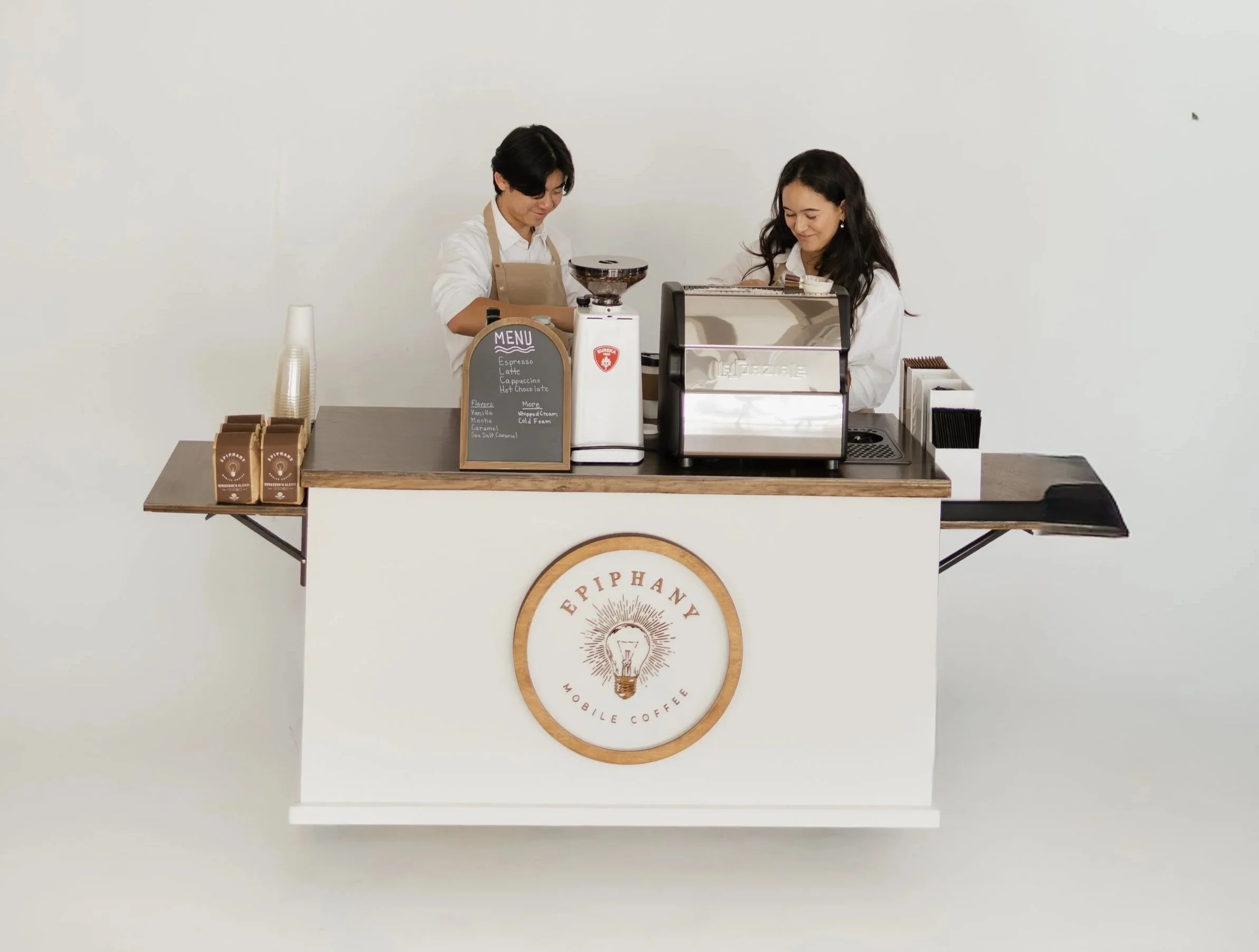 Two women working behind a white coffee counter with a wooden top, branded with 'Epipphany Mobile Coffee,' in a minimalist space, preparing coffee with a coffee grinder and an espresso machine.