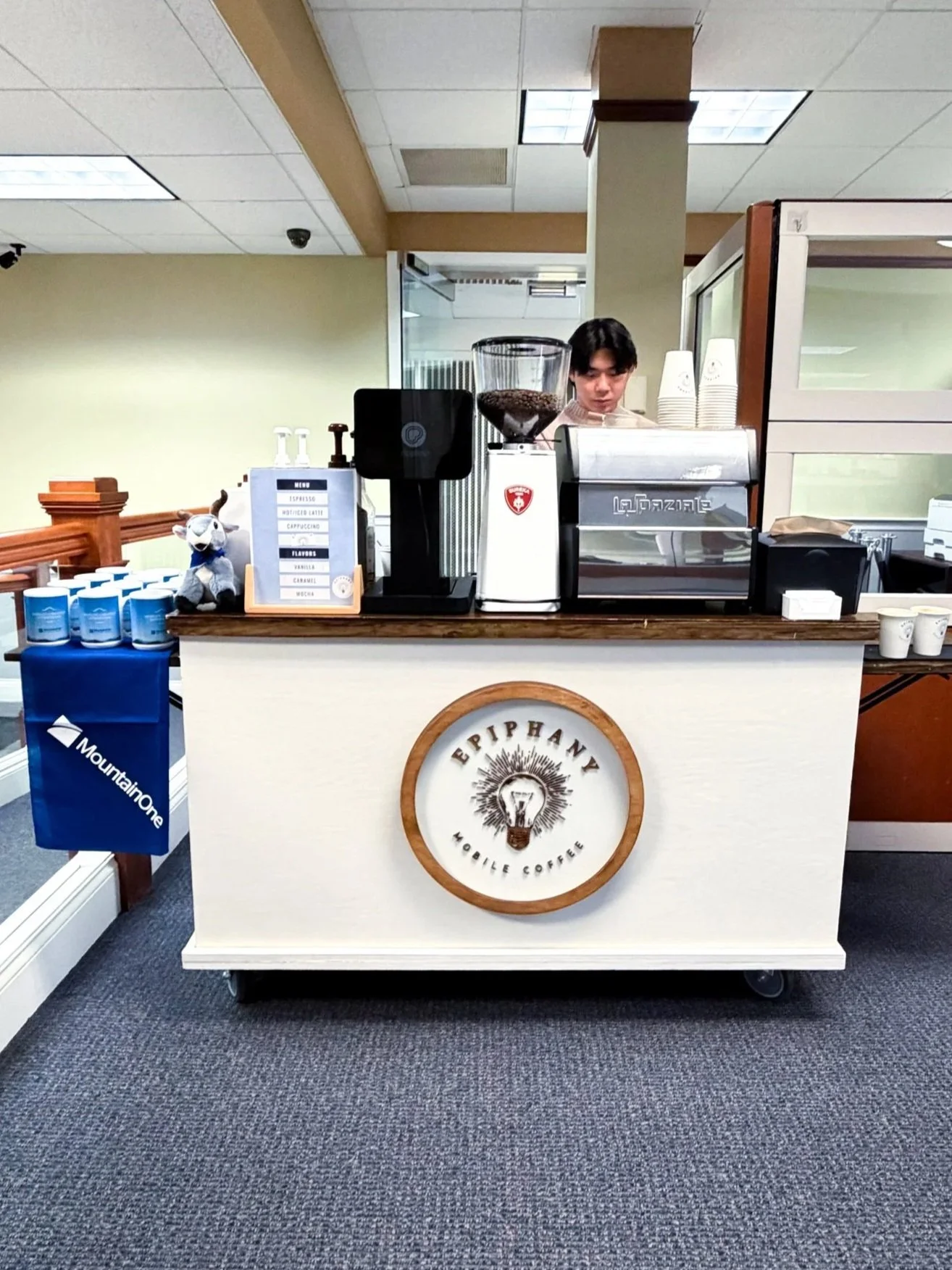 A coffee stand with a young barista behind it, featuring a logo that reads 'Epiphany Mobile Coffee' with a light bulb design. The stand also displays a menu, coffee cups, and brewing equipment, situated in an indoor setting with ceiling lights and beige walls.