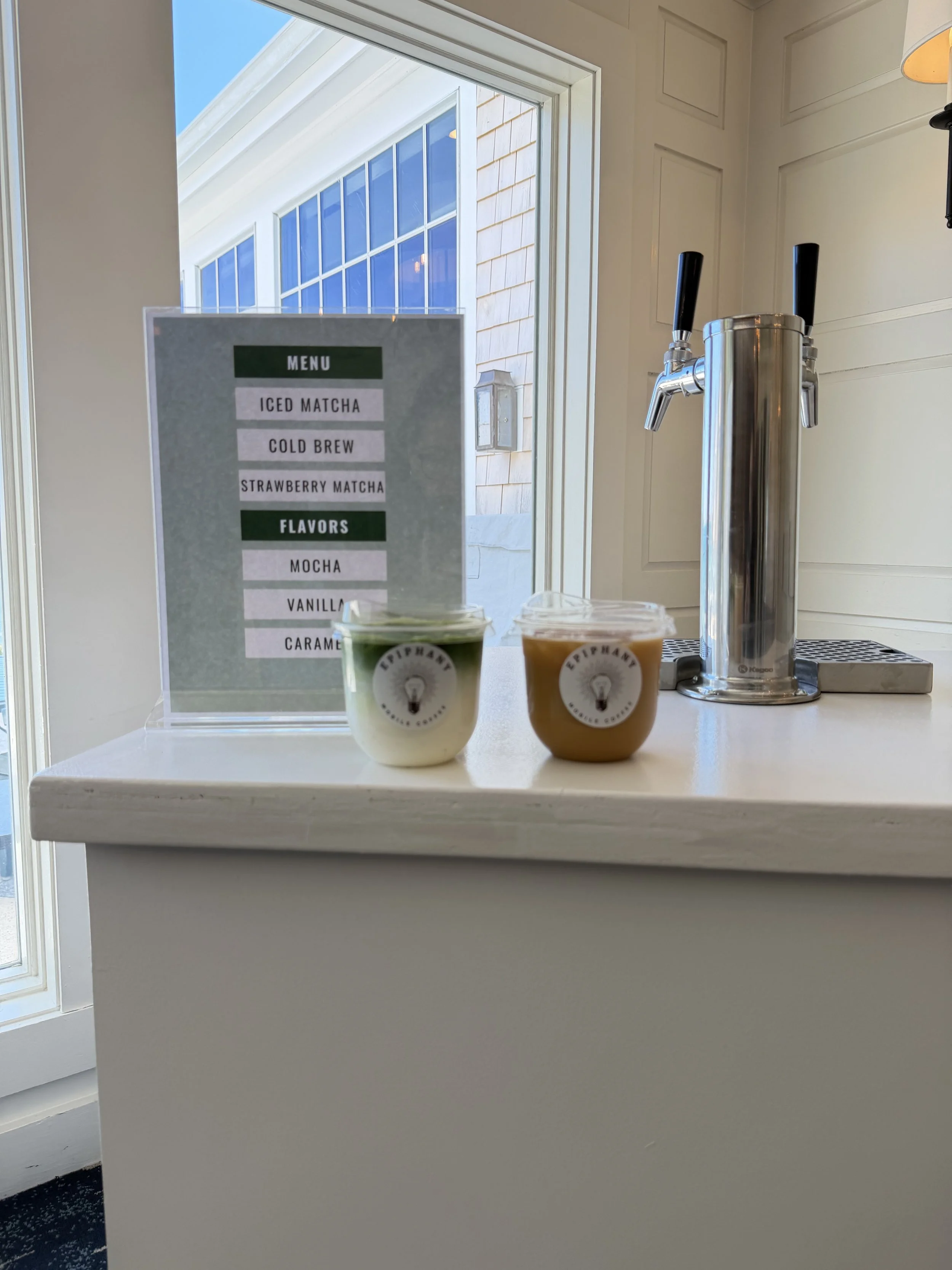 Interior view of a counter with two cups of matcha and caramel drinks, a menu sign listing iced matcha, cold brew, strawberry matcha, mocha, vanilla, and caramel flavors, a window with a blue sky outside, and a soda tap on the counter.