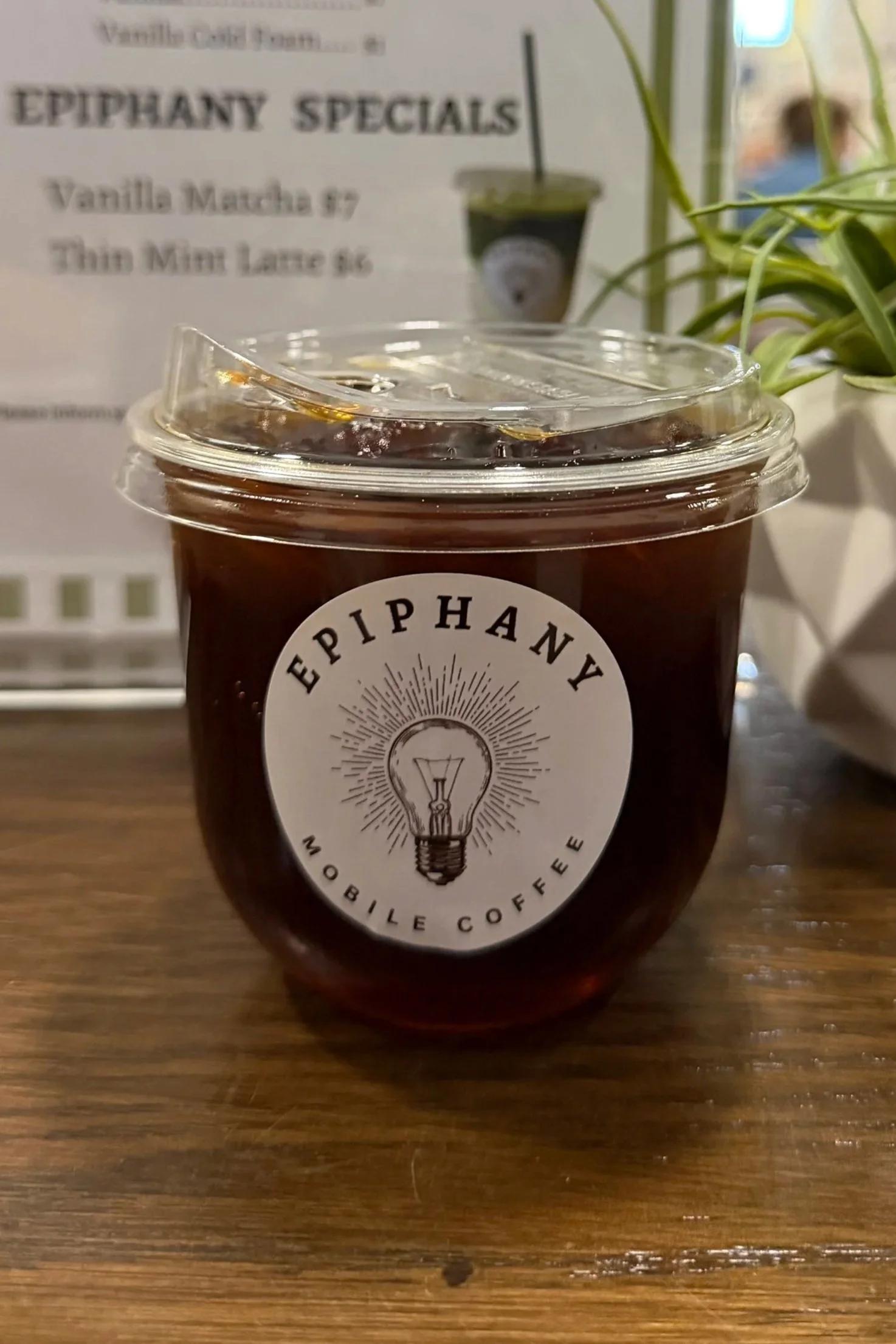 Close-up of a plastic cup filled with dark iced coffee or tea on a wooden table, with a lid and straw, featuring a logo of a light bulb with the words 'Epiphan' and 'Mobile Coffee' around it, and a blurred background of a menu sign and potted plant.