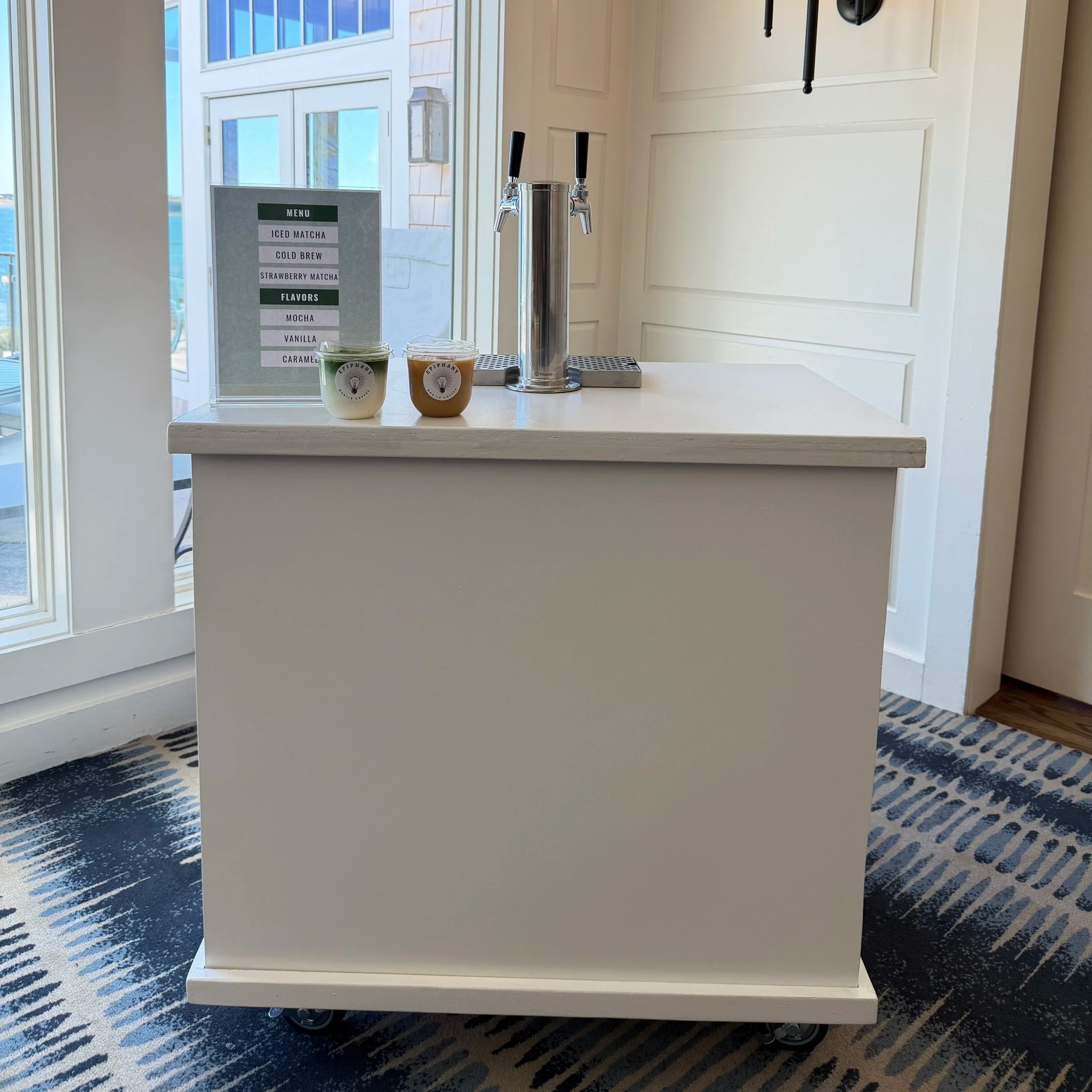 A white beverage cart on wheels with a tap system for dispensing drinks, placed on a patterned carpet near large windows. There is a menu sign in the background listing different drink options.