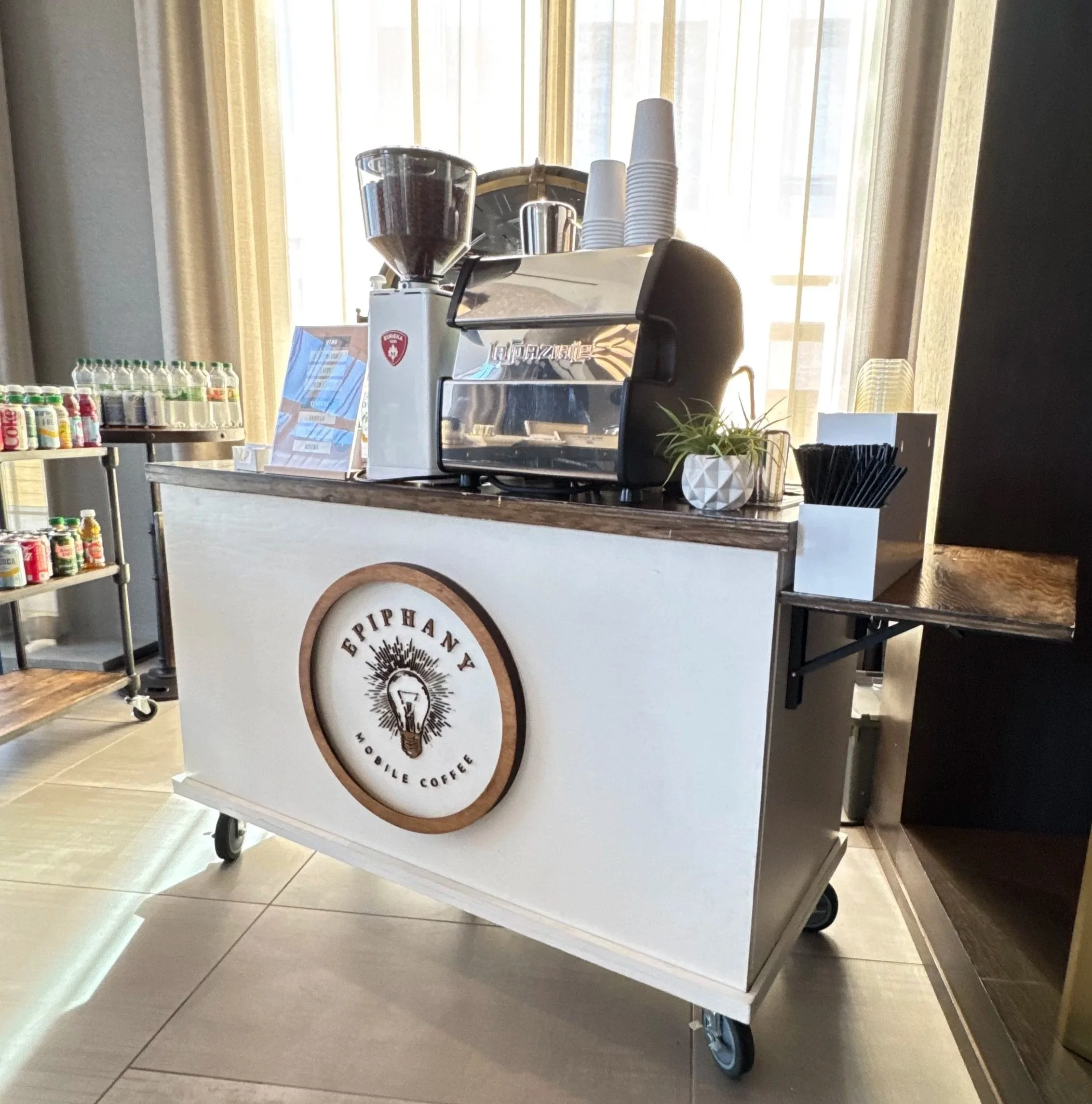 Coffee machine on a wooden counter with disposable cups stacked beside it. A metal container is also on the counter. To the left, a round display shelf holds small bottled drinks, and a sign or menu is partially visible. The background features large