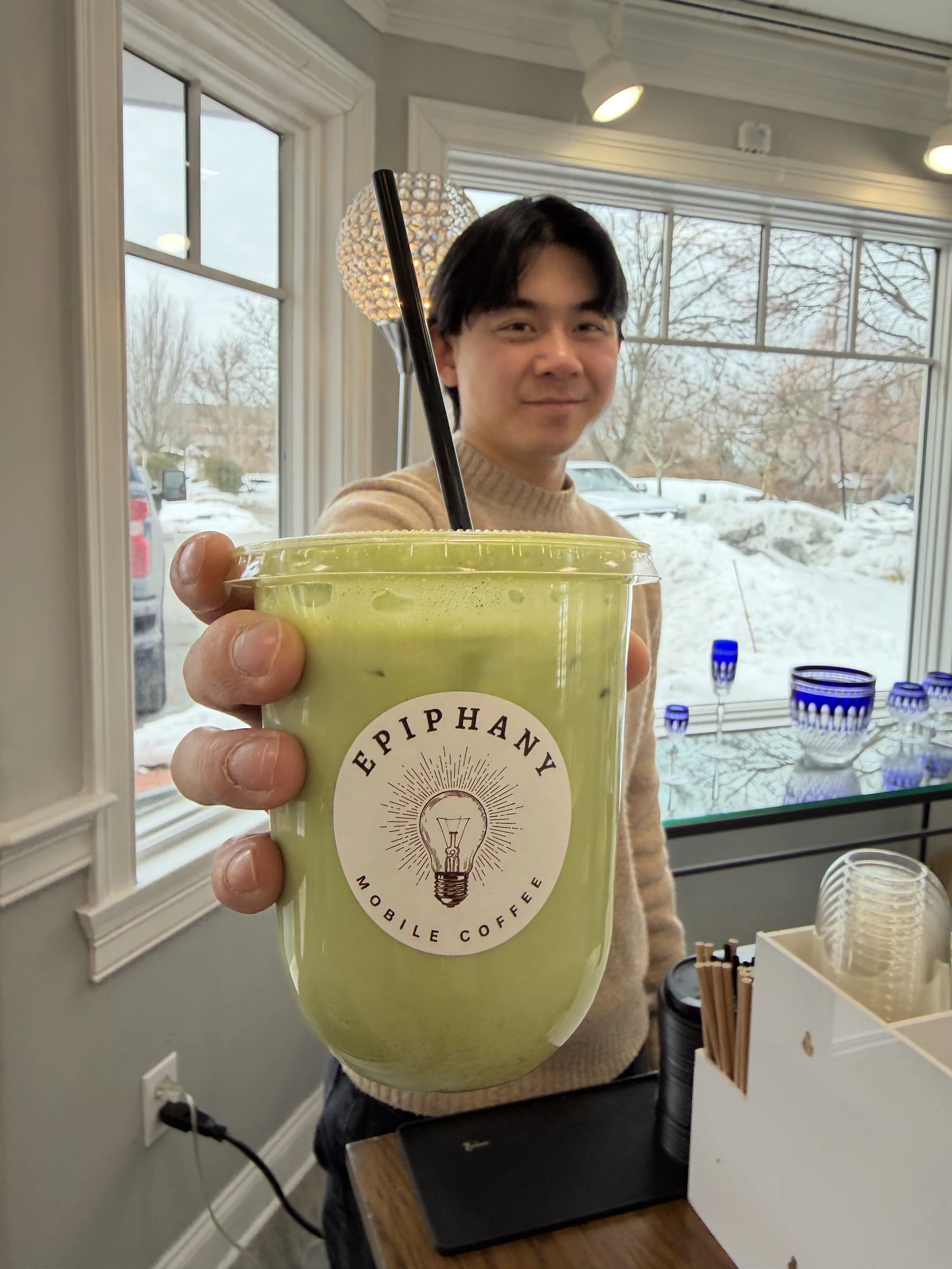A person holding a green cold beverage cup with a black straw, inside a room with large windows showing a snowy outdoor landscape and trees, with a lamp and table in the background.