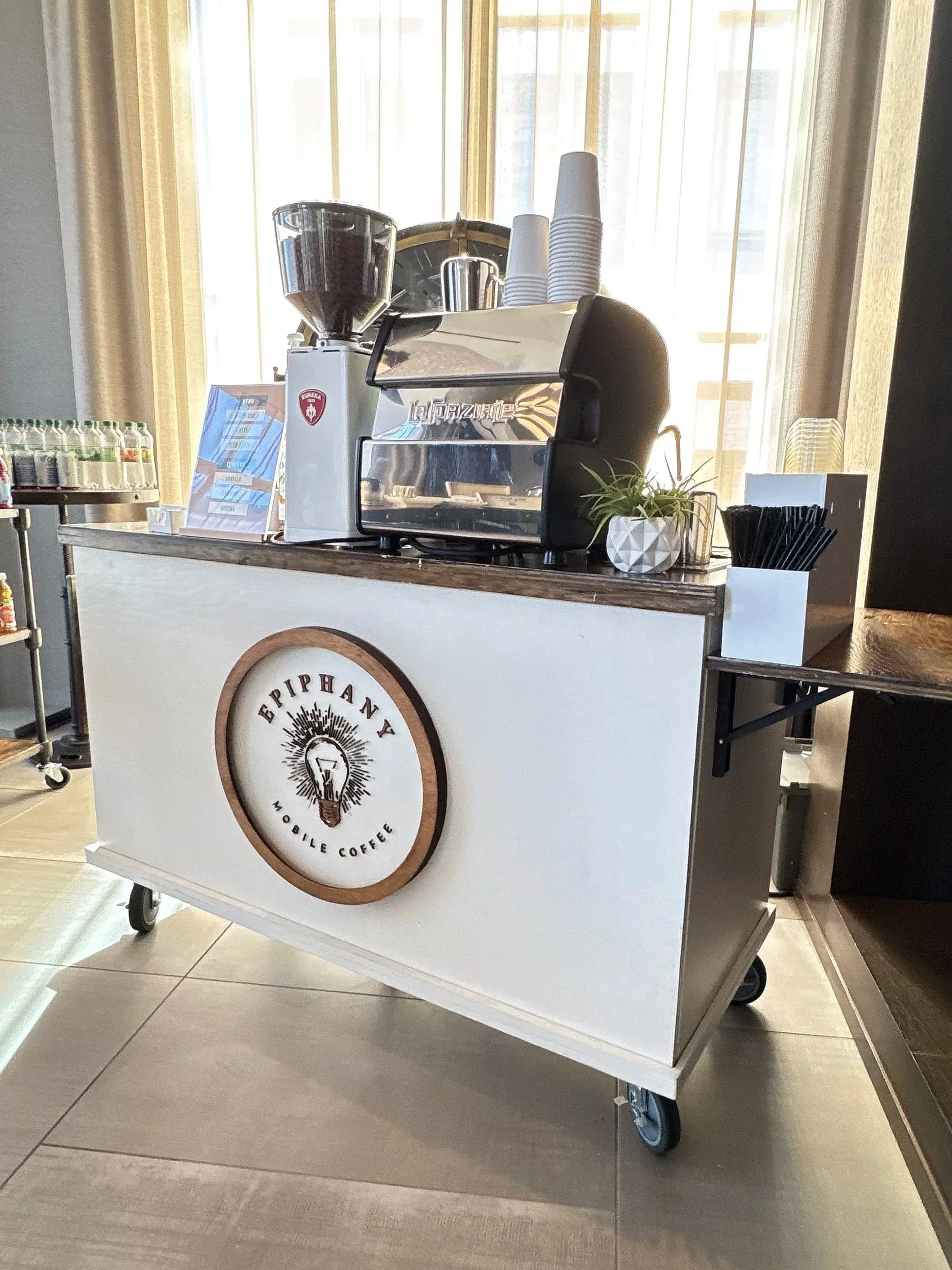 Mobile coffee cart with a sign that says 'Epiphany Mobile Coffee' on the front, displaying a coffee grinder, disposable cups, and a small plant, set near a window with curtains.