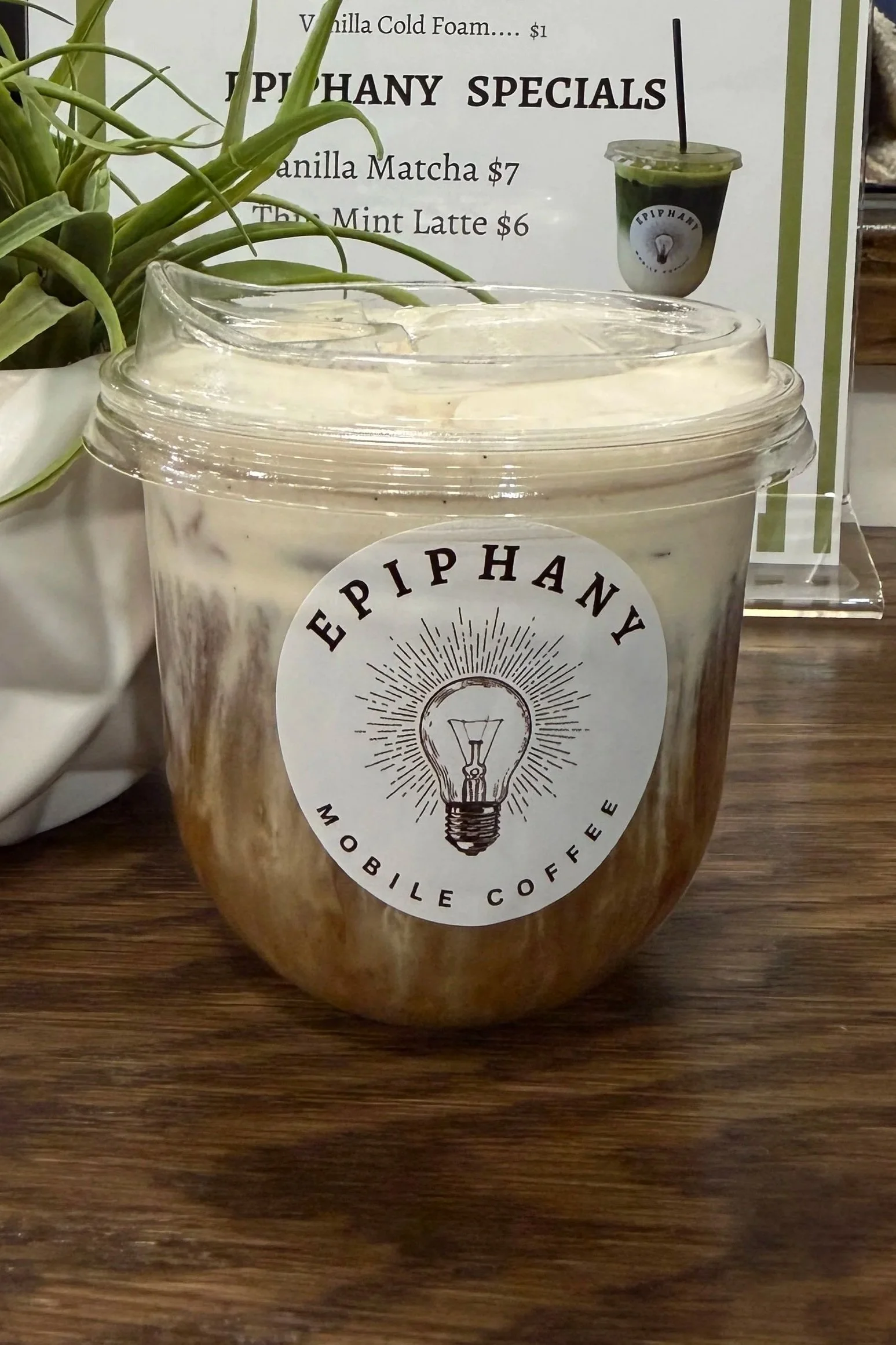 A clear plastic cooler with a lid containing iced coffee and a white sticker with the Eiphany logo, next to a potted plant and a menu board listing specialty drinks.