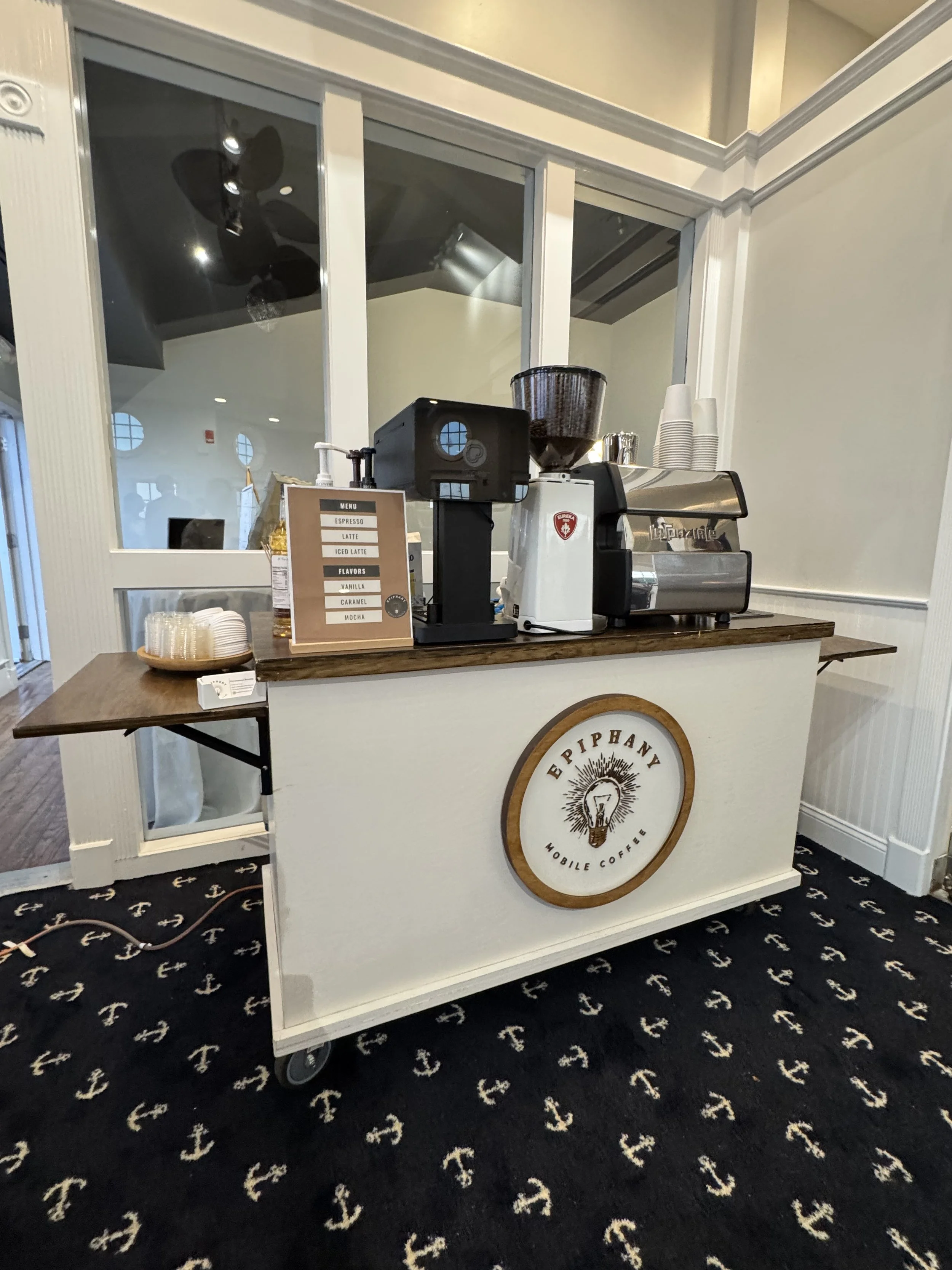 A mobile coffee cart with a sign listing espresso, latte, iced latte, vanilla, caramel, and mocha drinks. The cart has a coffee machine, cups, and a small tray of cups on top, with a logo that reads "Epphany Mobile Coffee." The background includes a 