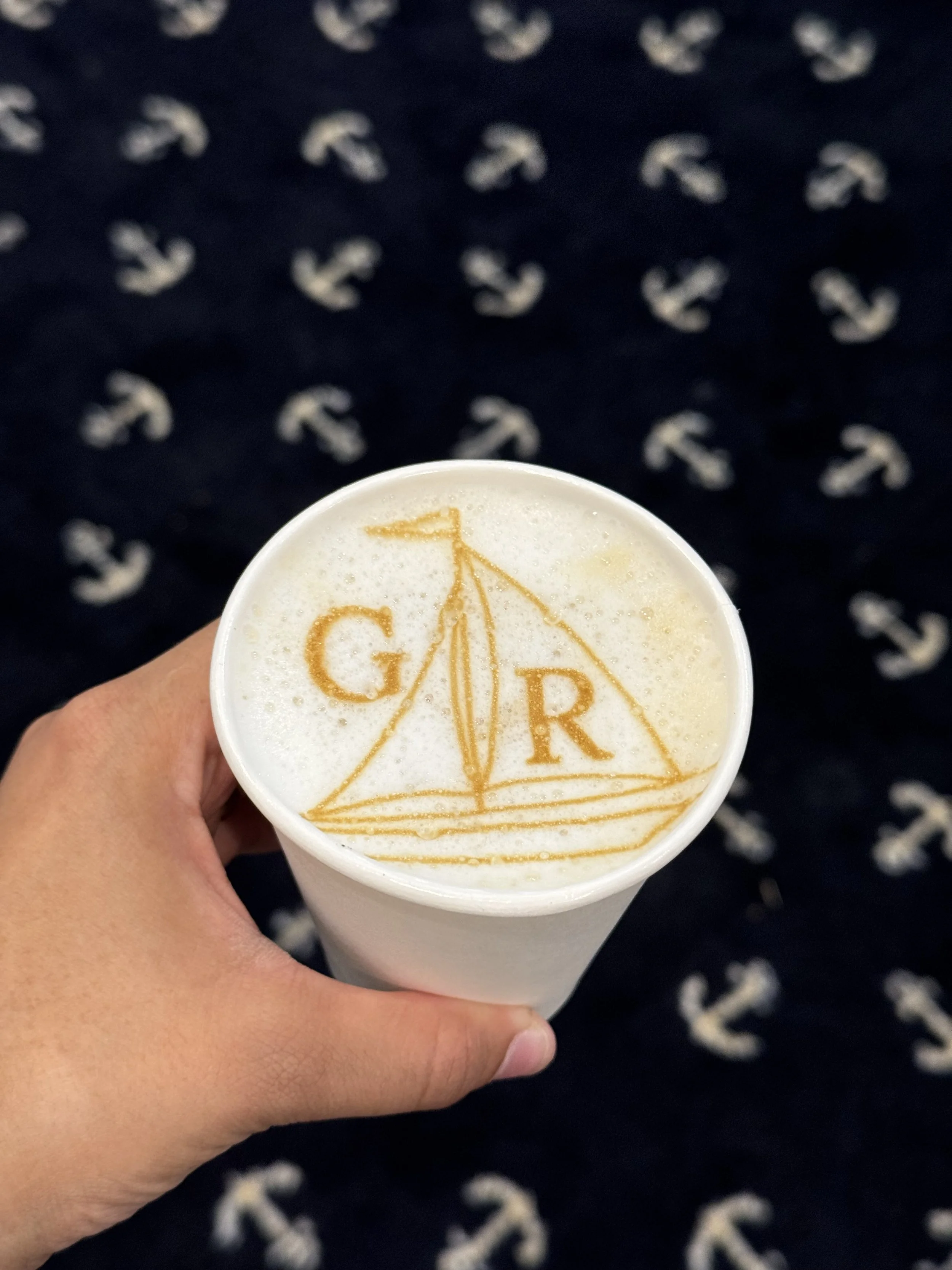 A hand holding a white cup with a latte displaying the letters G and R in caramel on the foam, with an anchor-patterned fabric background.