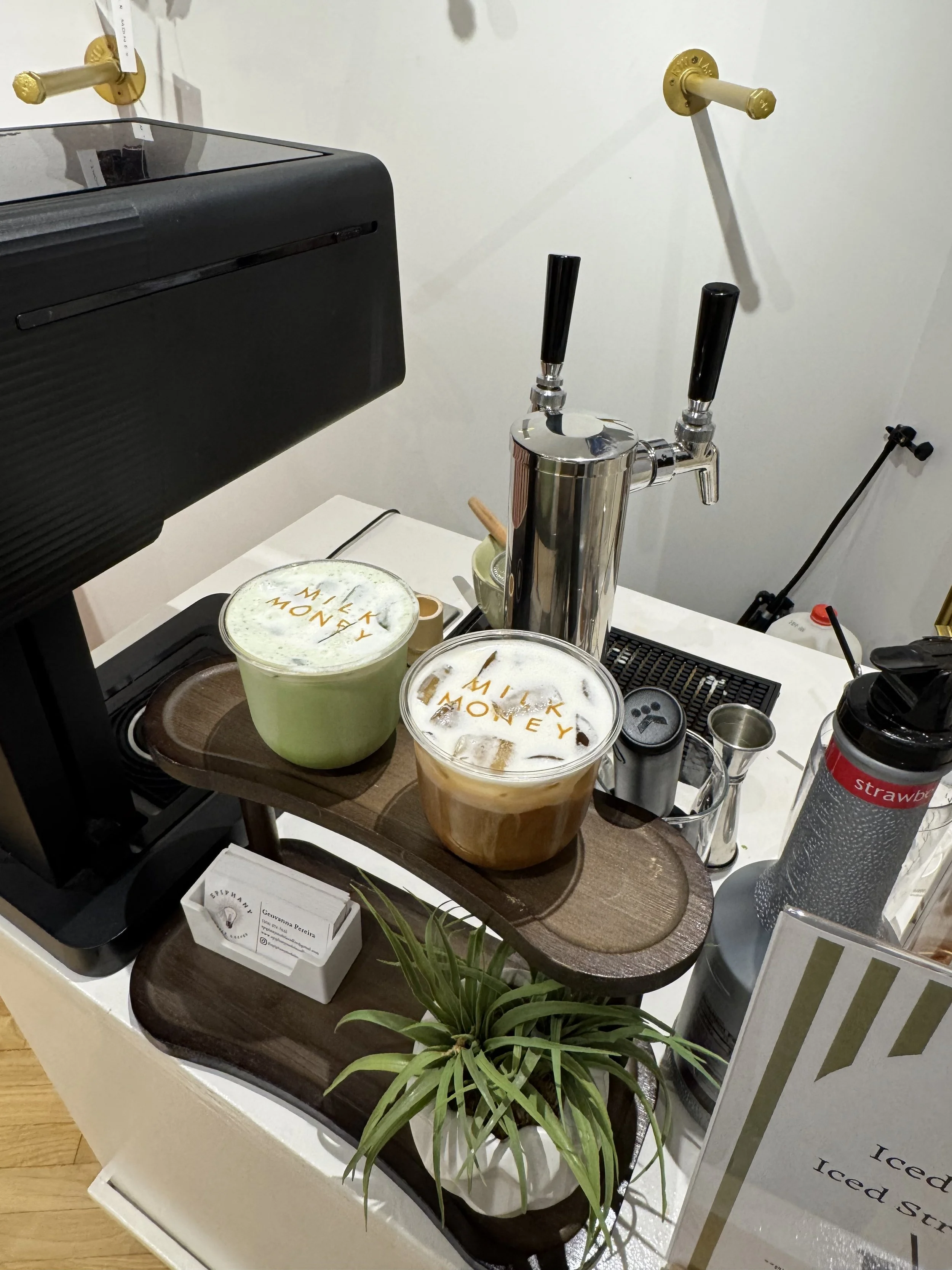 Two cups of coffee with foam and the words 'Milk Money' written on the foam, placed on a wooden tray on a white table, with a black coffee machine, green plant, and a small box of business cards nearby.