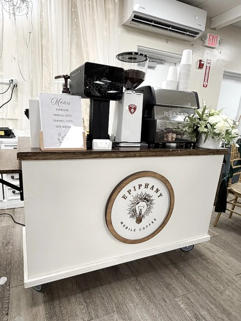 A mobile coffee stand with a sign displaying the coffee menu, a coffee grinder, an espresso machine, paper cups, and a vase with white flowers. The stand has a circular logo reading 'Epiphany Mobile Coffee' with a light bulb illustration.