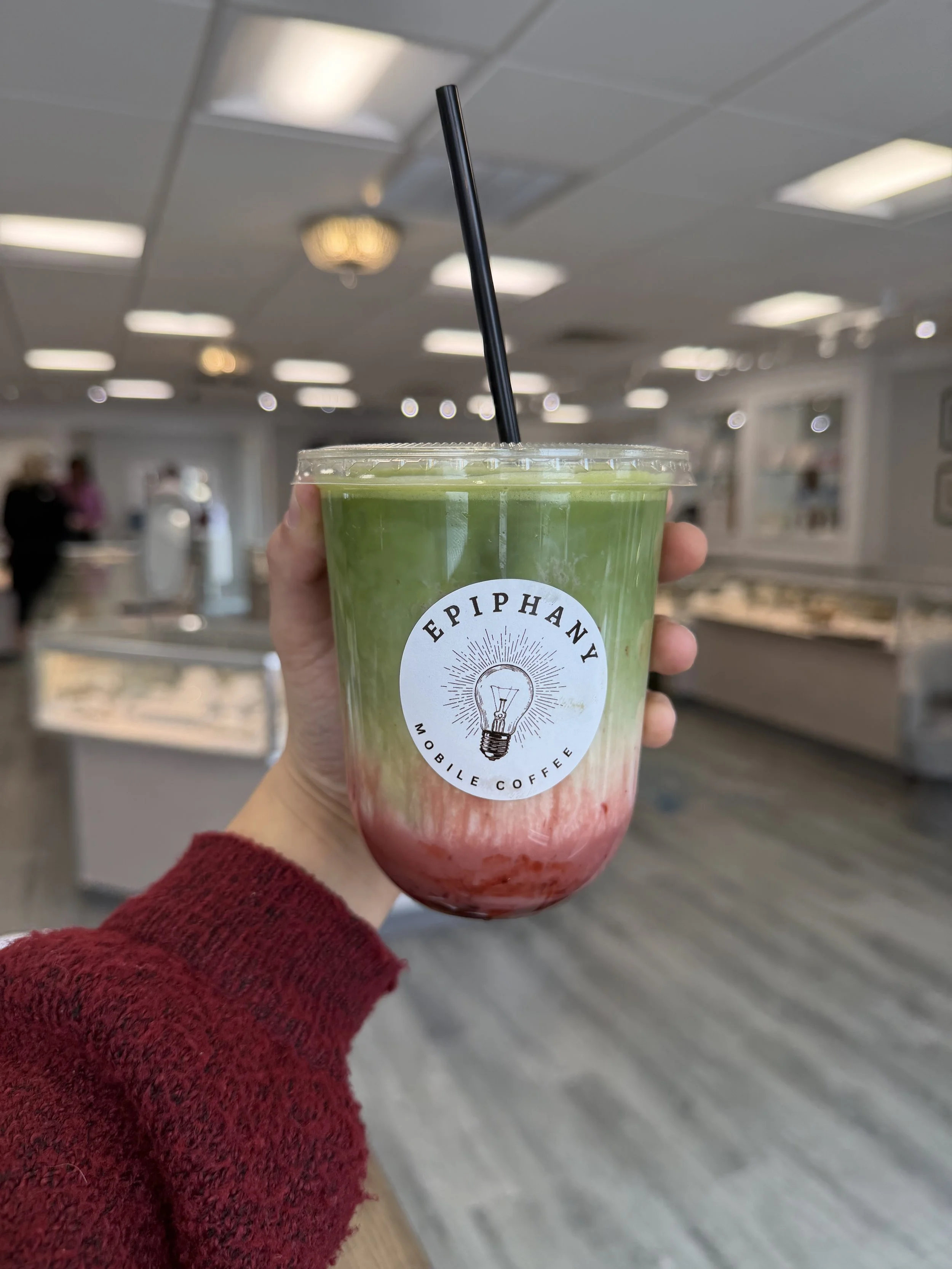Hand holding a clear plastic cup with green and red layered smoothie, with logo reading 'Eccentric Mobile Coffee' and a lightbulb graphic, in a room with ceiling lights and blurred background.