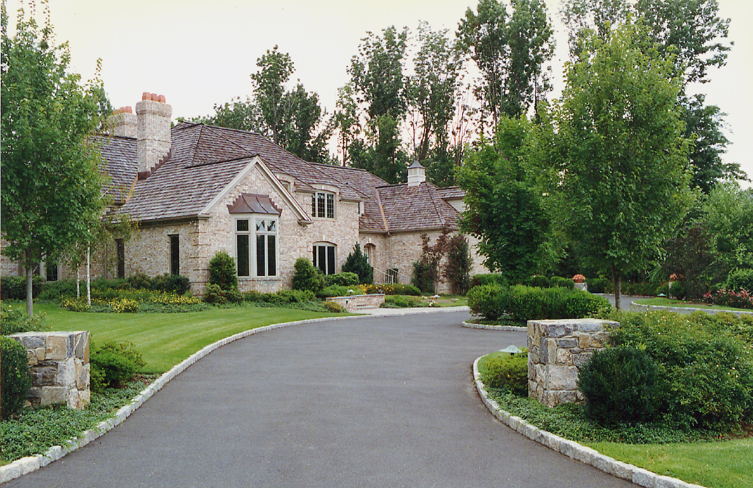 A large brick house with a landscaped front yard and a curved asphalt driveway, surrounded by green trees and bushes.