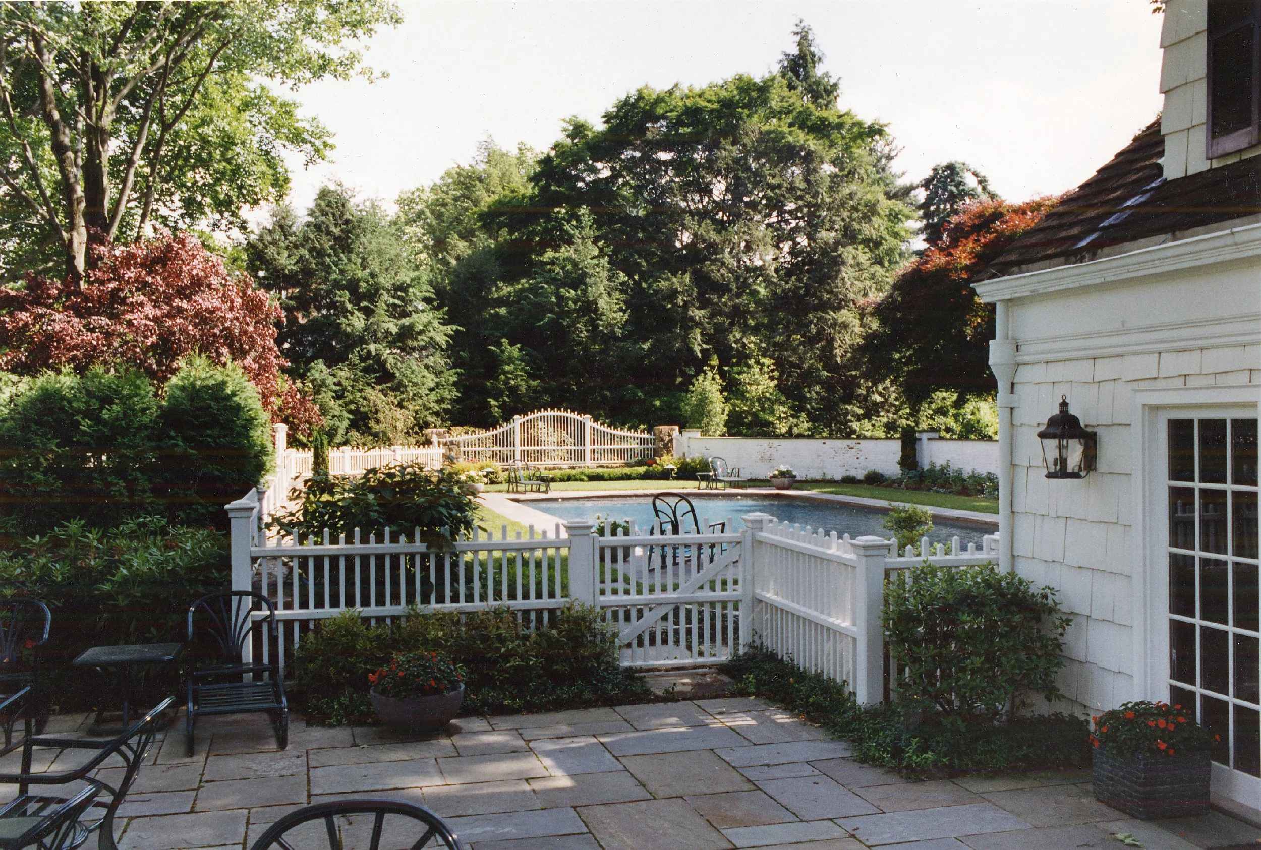 A backyard with a patio, white fence and gate, a pool, trees, shrubs, and a house with a lantern outside one window.
