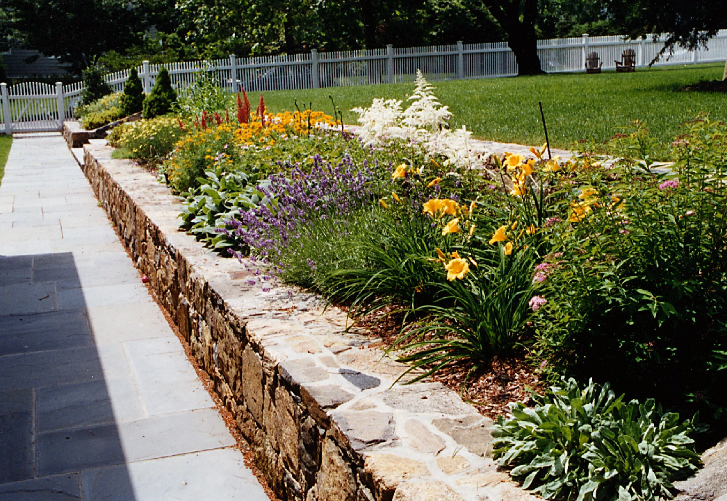 Colorful flower garden with a stone border, green lawn, and white picket fence in the background.