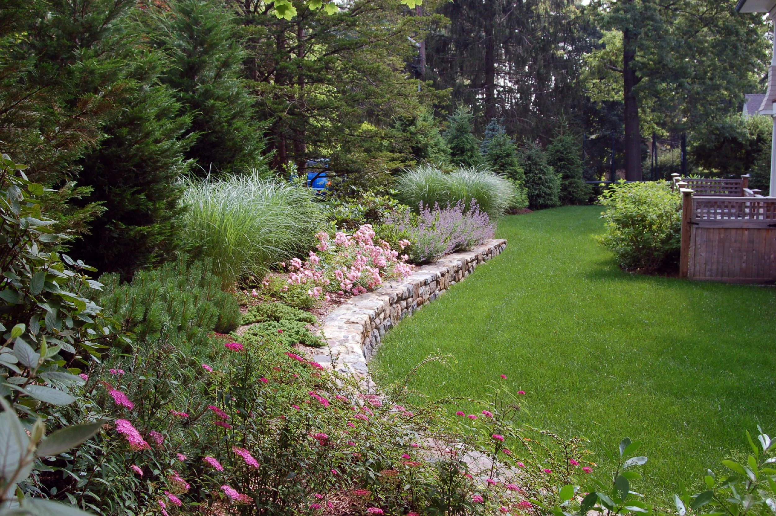 Lush green backyard with a well-maintained lawn, a stone-edged flower bed filled with pink and purple flowers, and a wooden fence in the background.