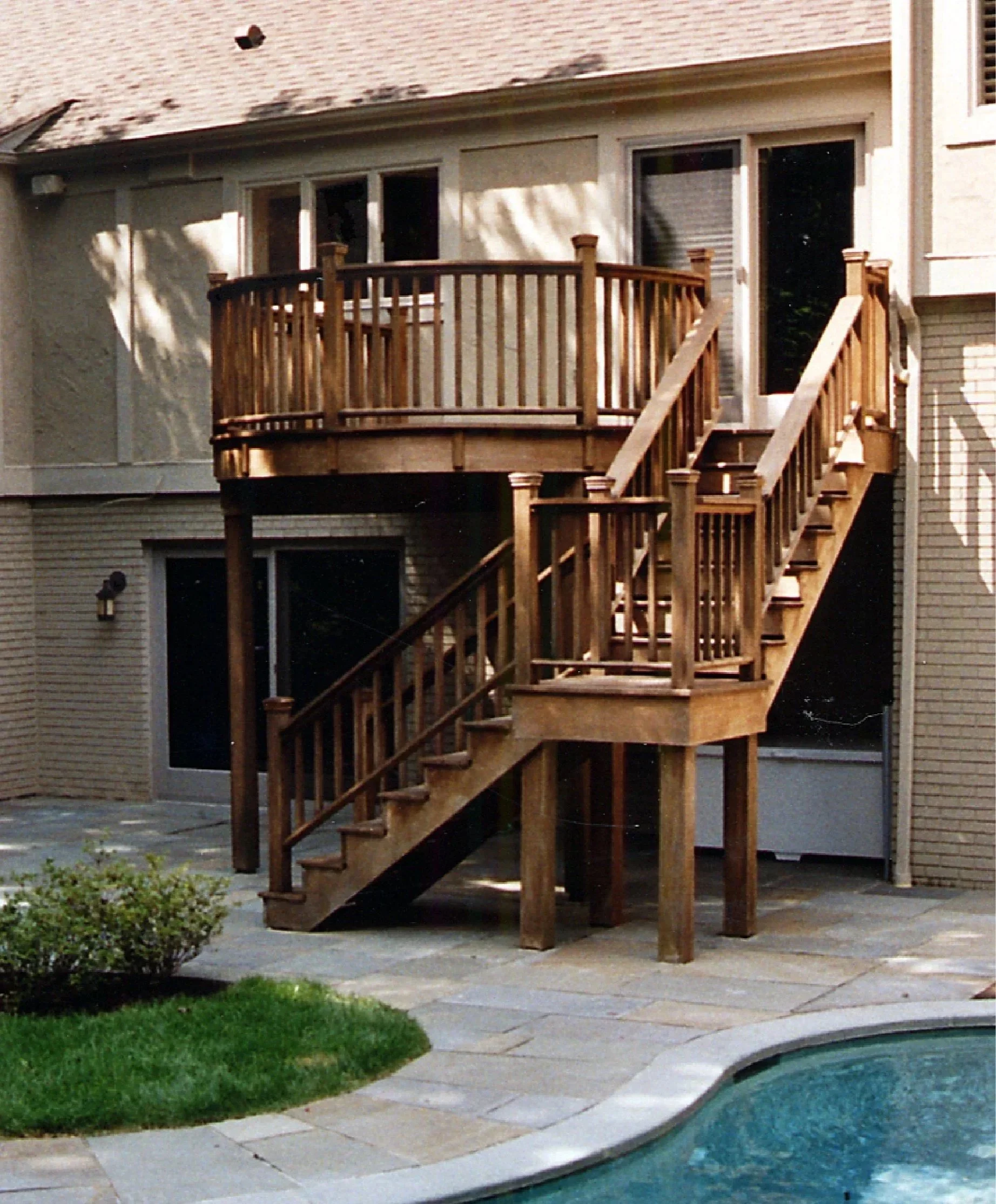 Wooden deck with stairs leading to a backyard with a swimming pool and a bush in the foreground.