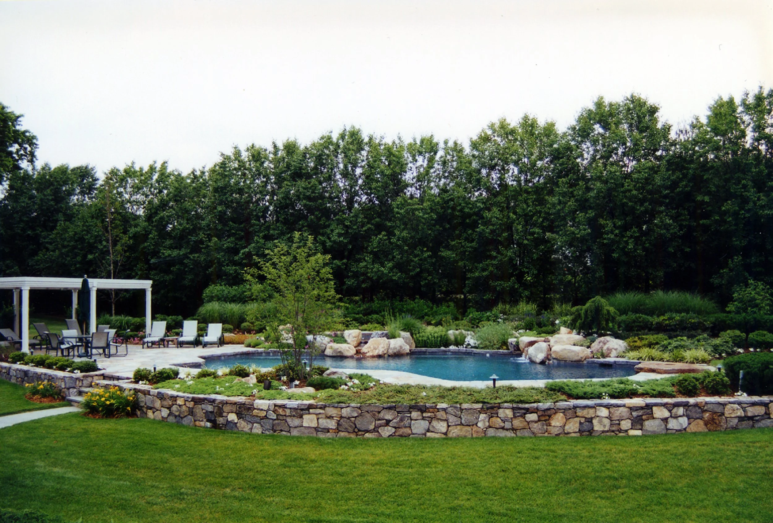 Backyard with a swimming pool surrounded by rocks, plants, and trees, with outdoor furniture and a covered patio area.