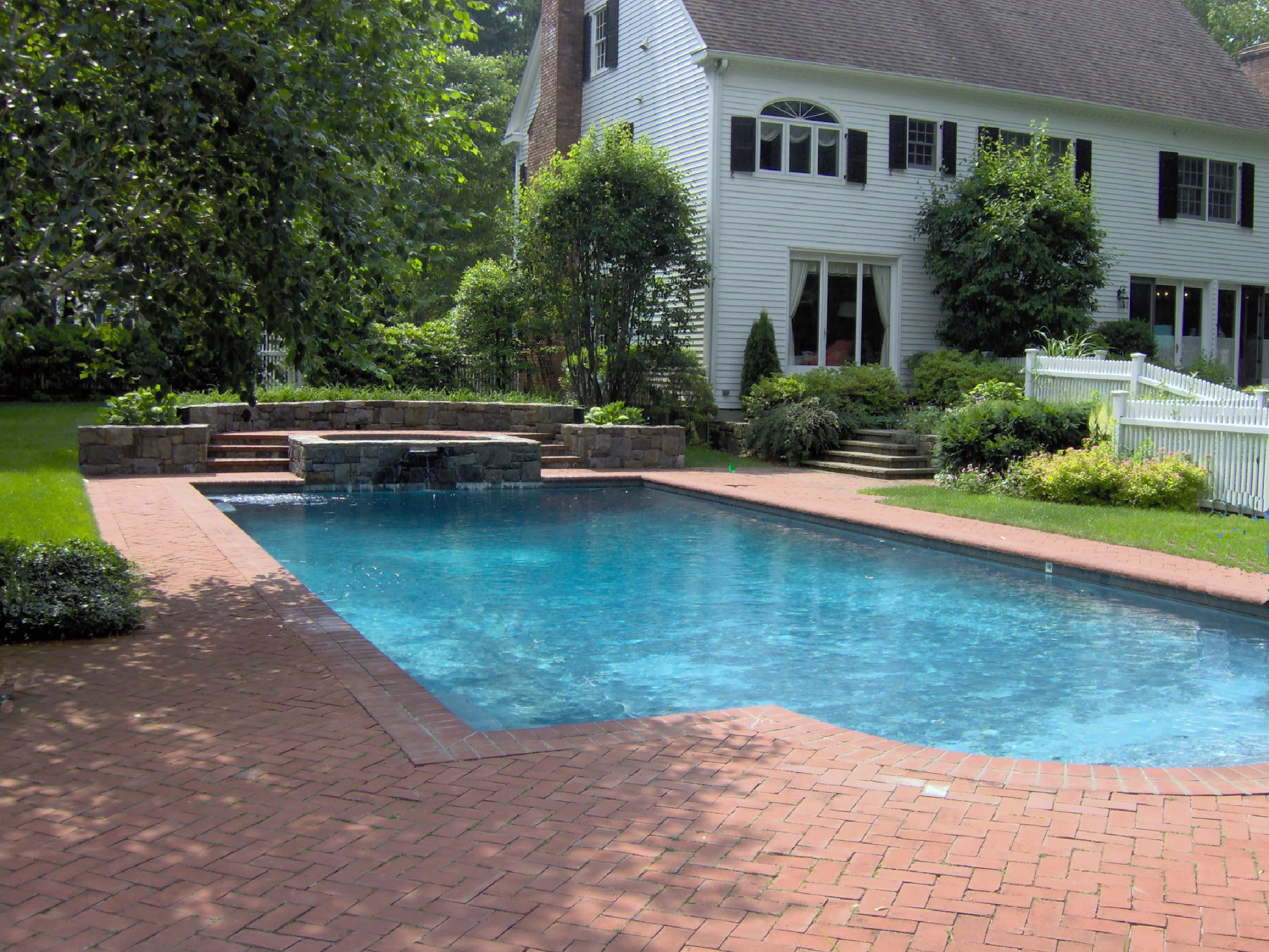 Residential backyard with in-ground swimming pool, brick patio, trees, bushes, and a white house with black shutters.
