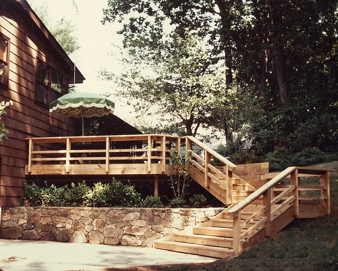 A newly built wooden staircase and deck attached to a house, with a green umbrella on the deck, surrounded by trees and landscaping stone wall.