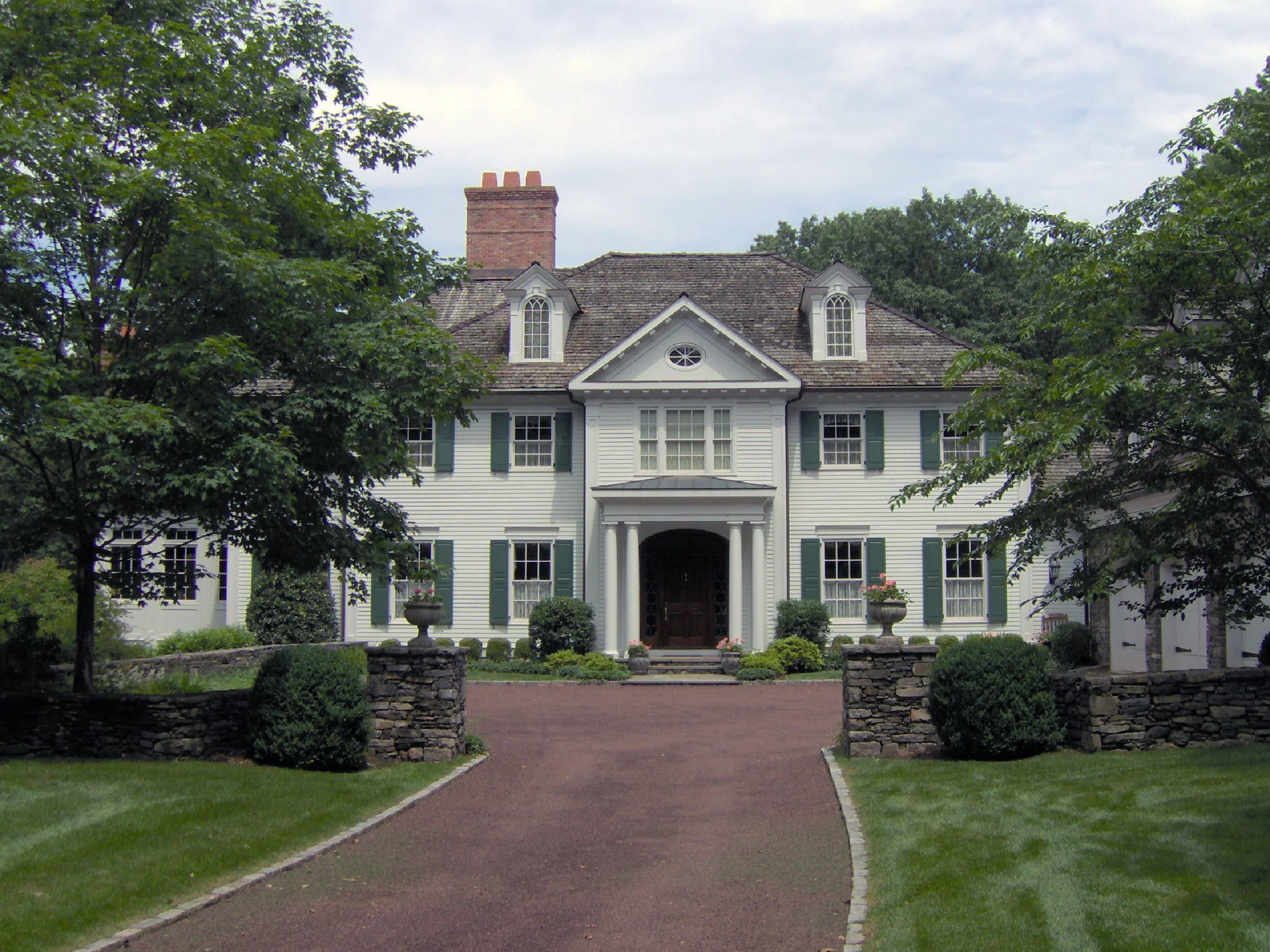 A large white house with green shutters, a brick driveway, trees, and a stone wall in front.