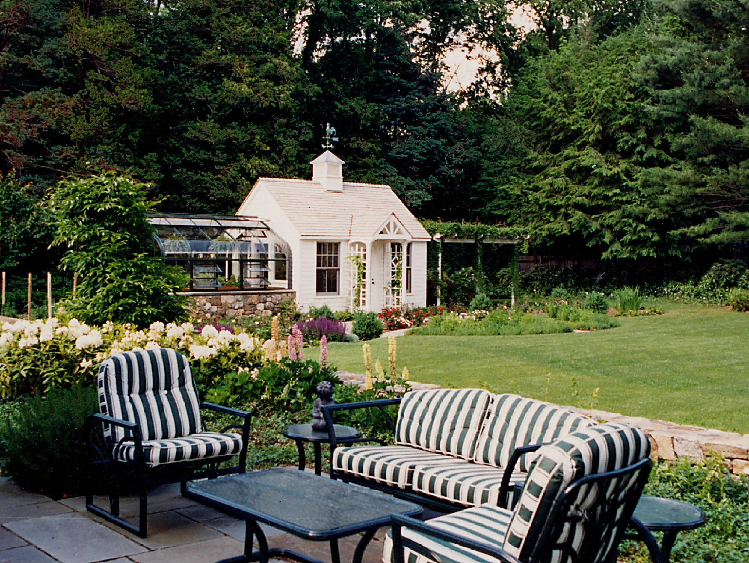 A backyard garden with striped outdoor furniture, a small white shed, a greenhouse, lush green trees, and a well-kept lawn.