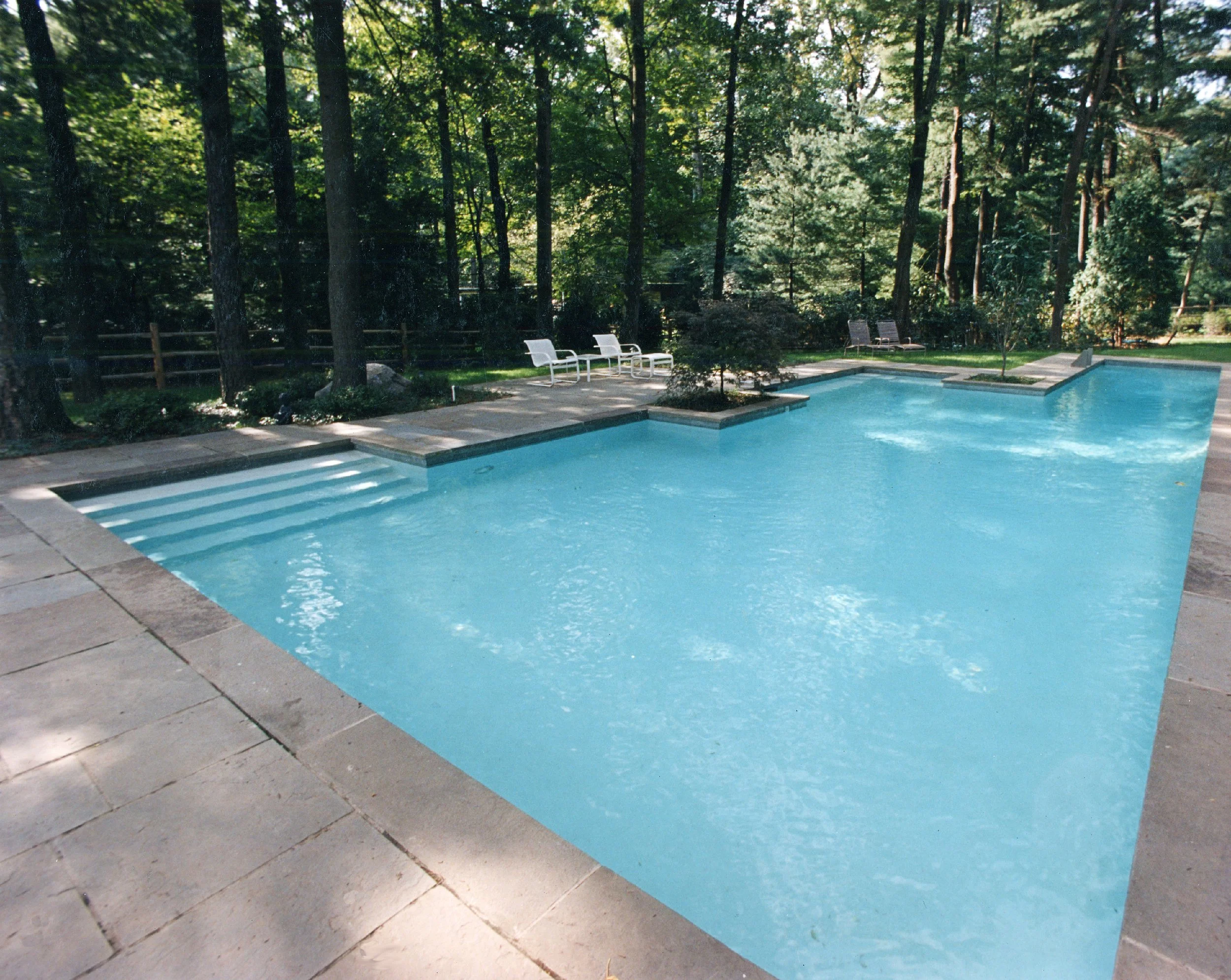 A backyard swimming pool surrounded by trees and lounge chairs.
