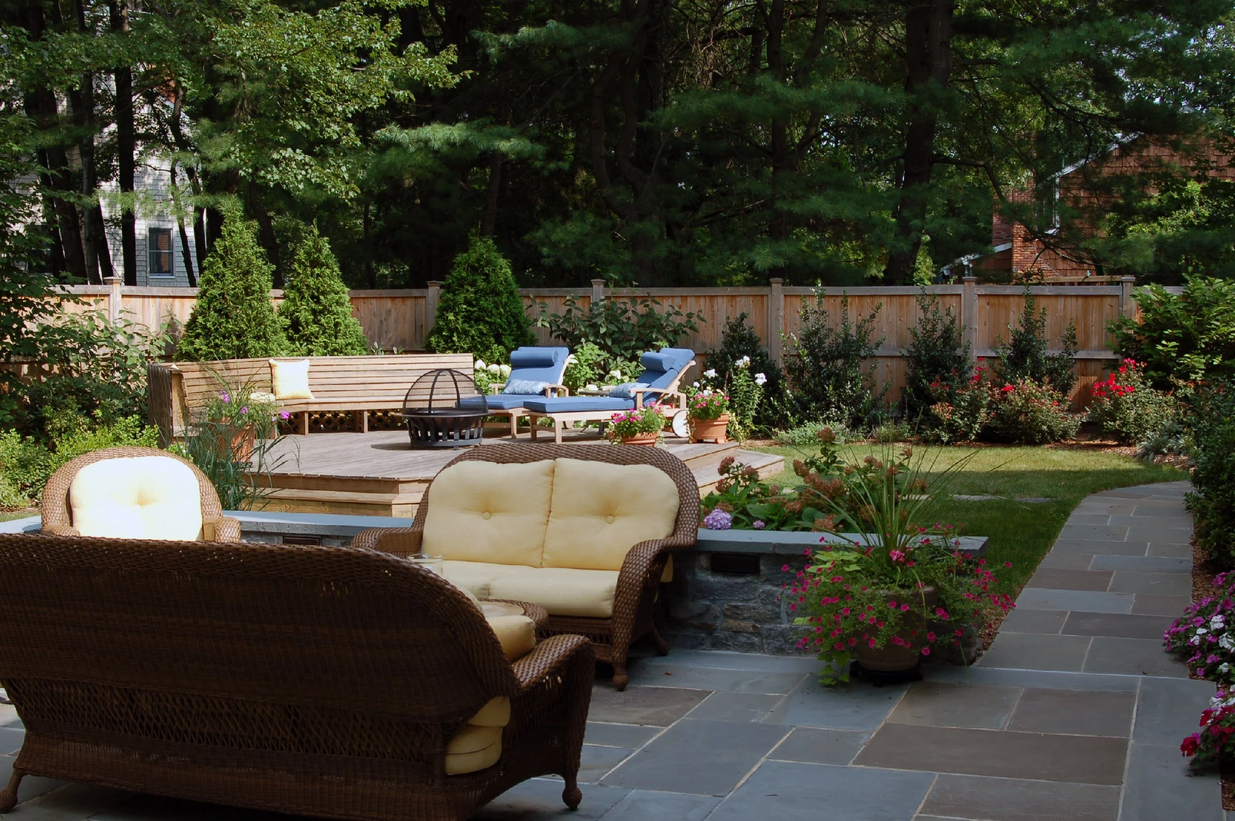 A backyard patio with wicker and cushioned outdoor furniture, flowering potted plants, a landscaped garden with bushes and trees, and a wooden fence in the background.