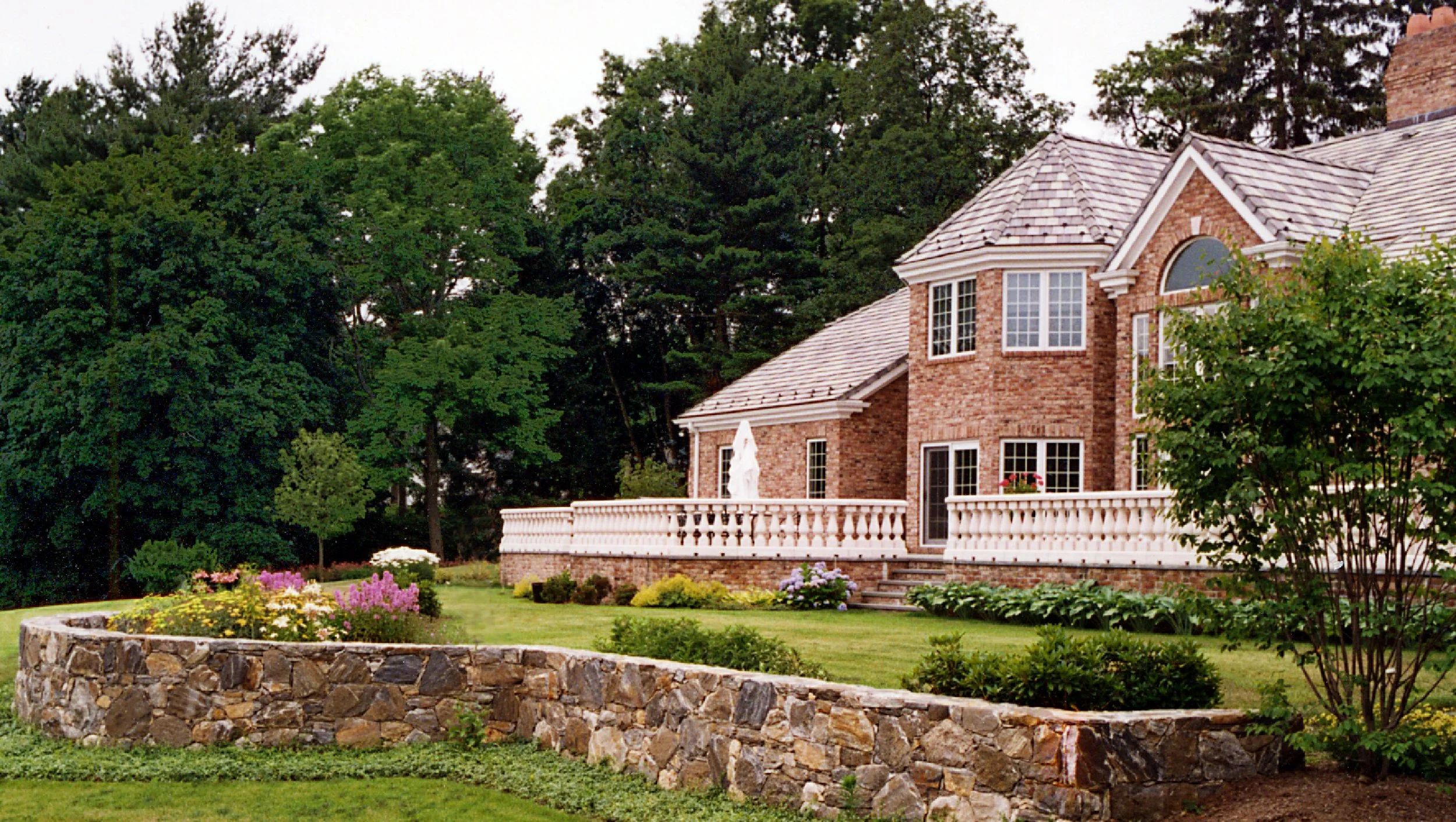 A large brick house with multiple windows and a balcony, surrounded by a well-maintained lawn and landscaped garden with colorful flowers, stone border, and tall trees in the background.