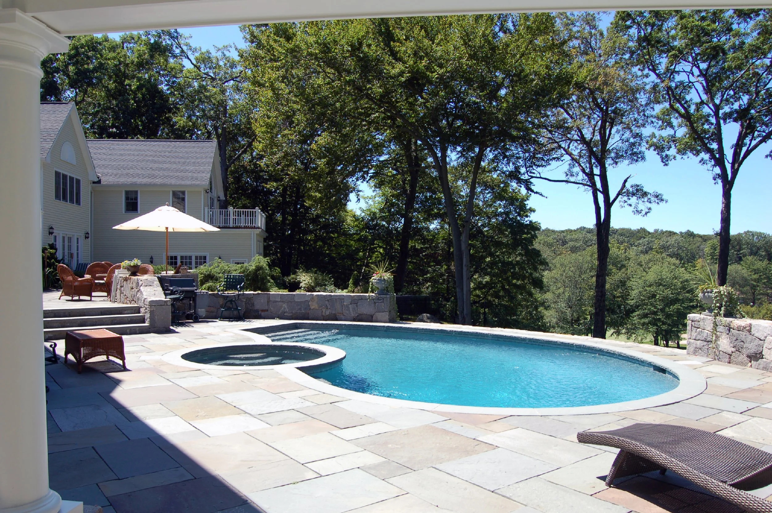 A backyard with a swimming pool, hot tub, outdoor furniture, umbrella, and trees in the background.